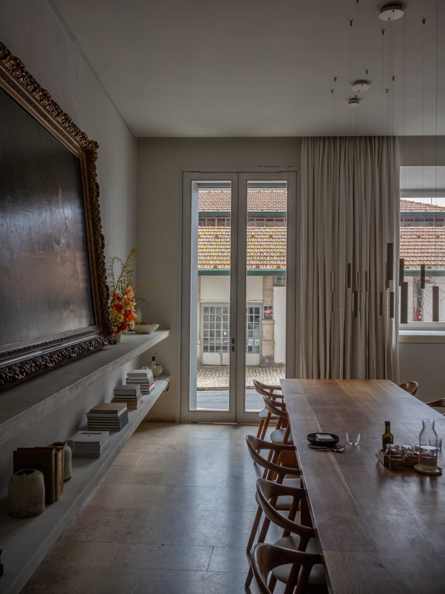 Santa Clara 1728, Portugal — Minimalist dining room with walnut table, mid-century chairs, ornate gold-framed artwork, terrazzo flooring, and floor-to-ceiling curtains overlooking historic tiled roofscape.