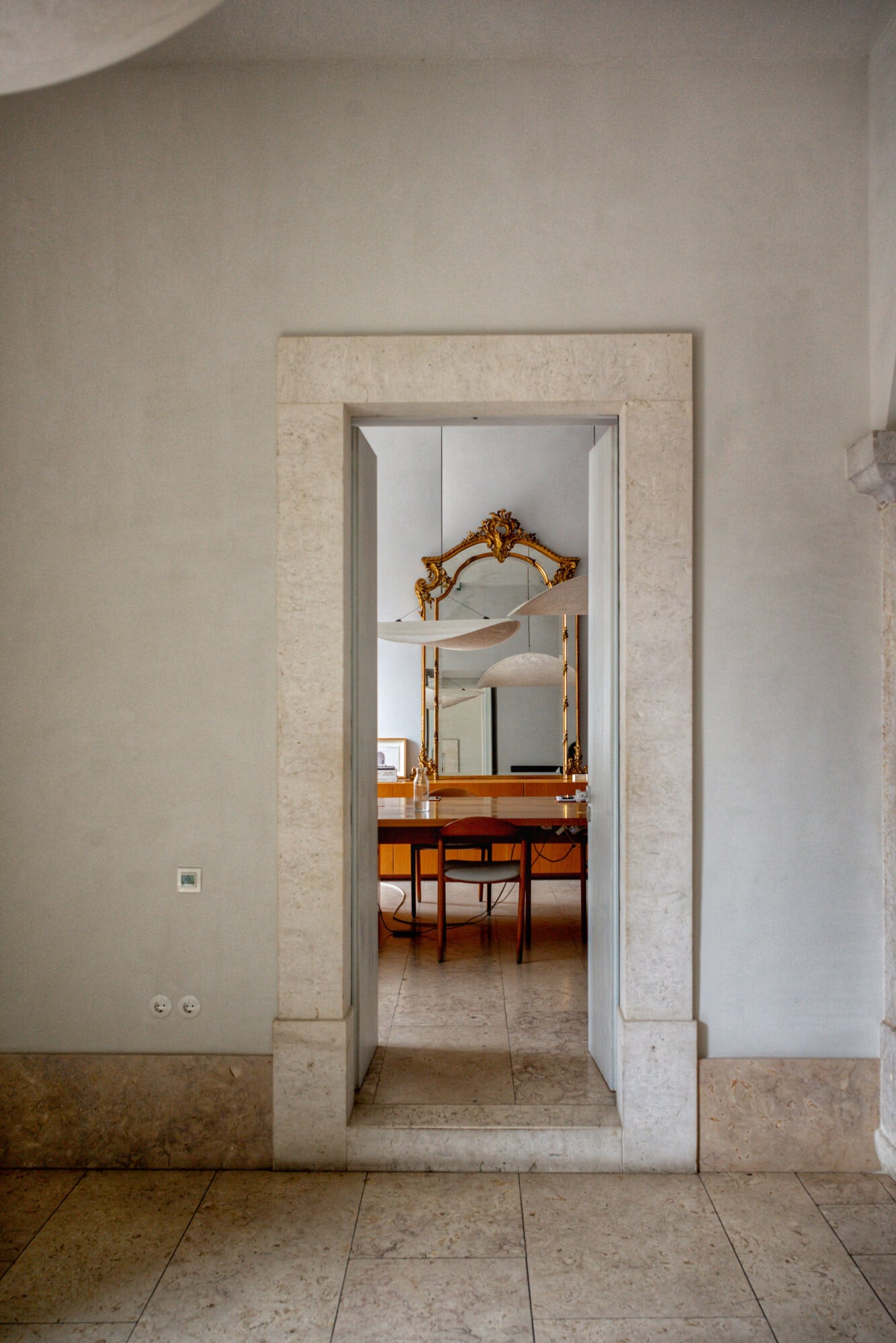 Santa Clara 1728, Portugal — Travertine-framed doorway opening to mid-century bedroom with gilt mirror and warm wood furnishings, minimalist contemporary interiors.