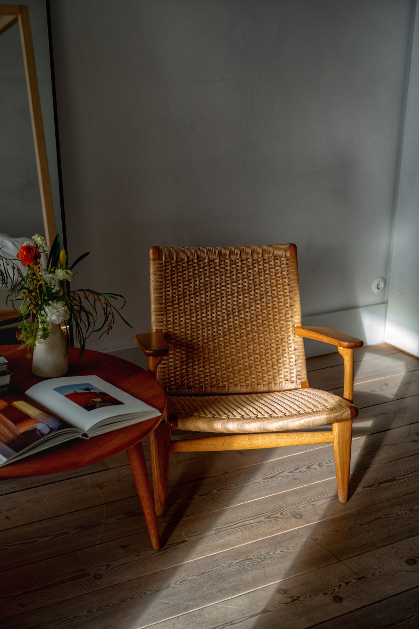 Santa Clara 1728, Portugal — Woven rattan lounge chair with wooden frame in sunlit room beside open book and fresh flowers on side table.