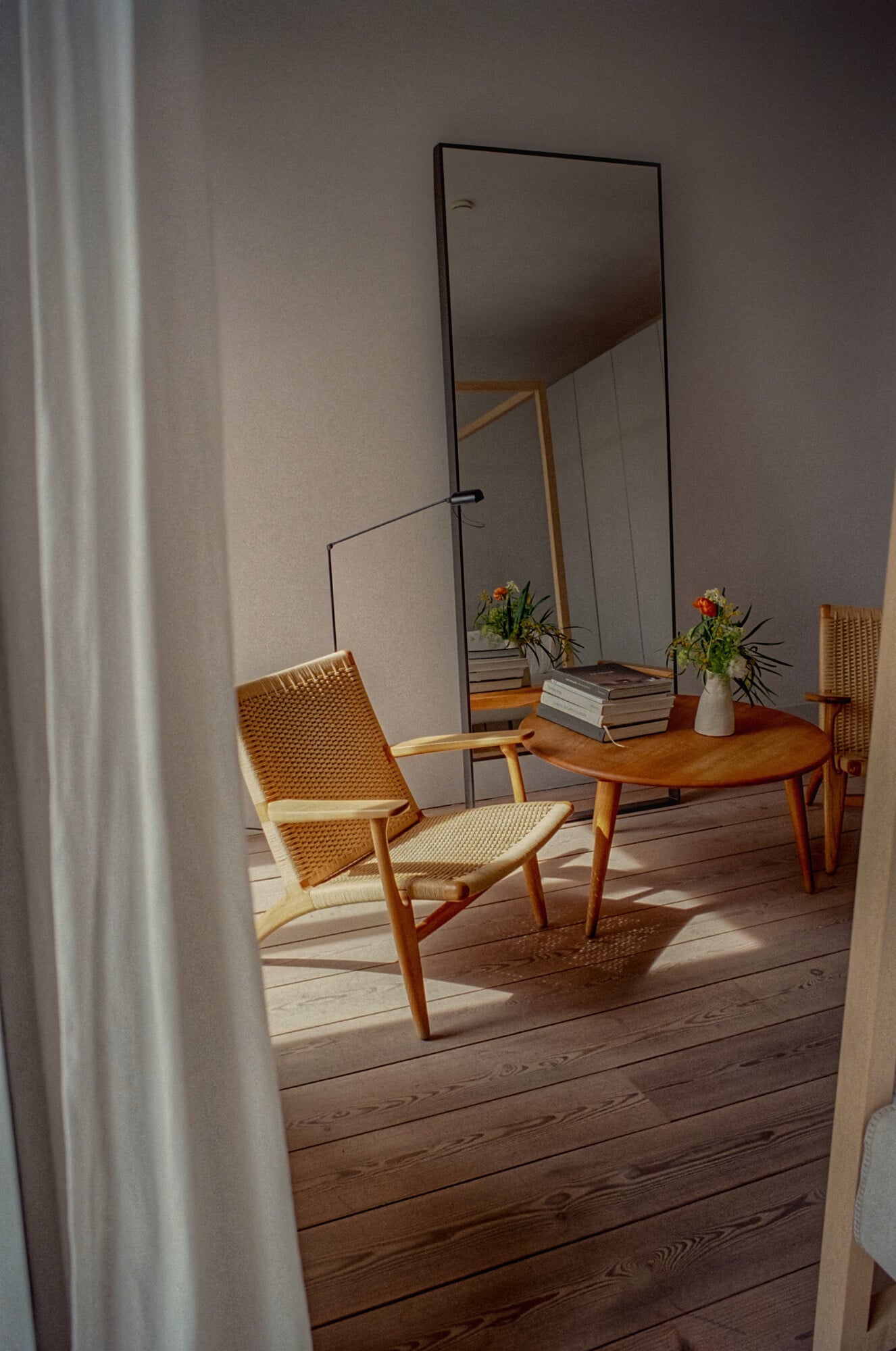 Santa Clara 1728, Portugal — Mid-century wooden chair with woven seat beside round table, tall mirror, fresh flowers, stacked books, warm sunlight on wooden floors, minimalist interior design.