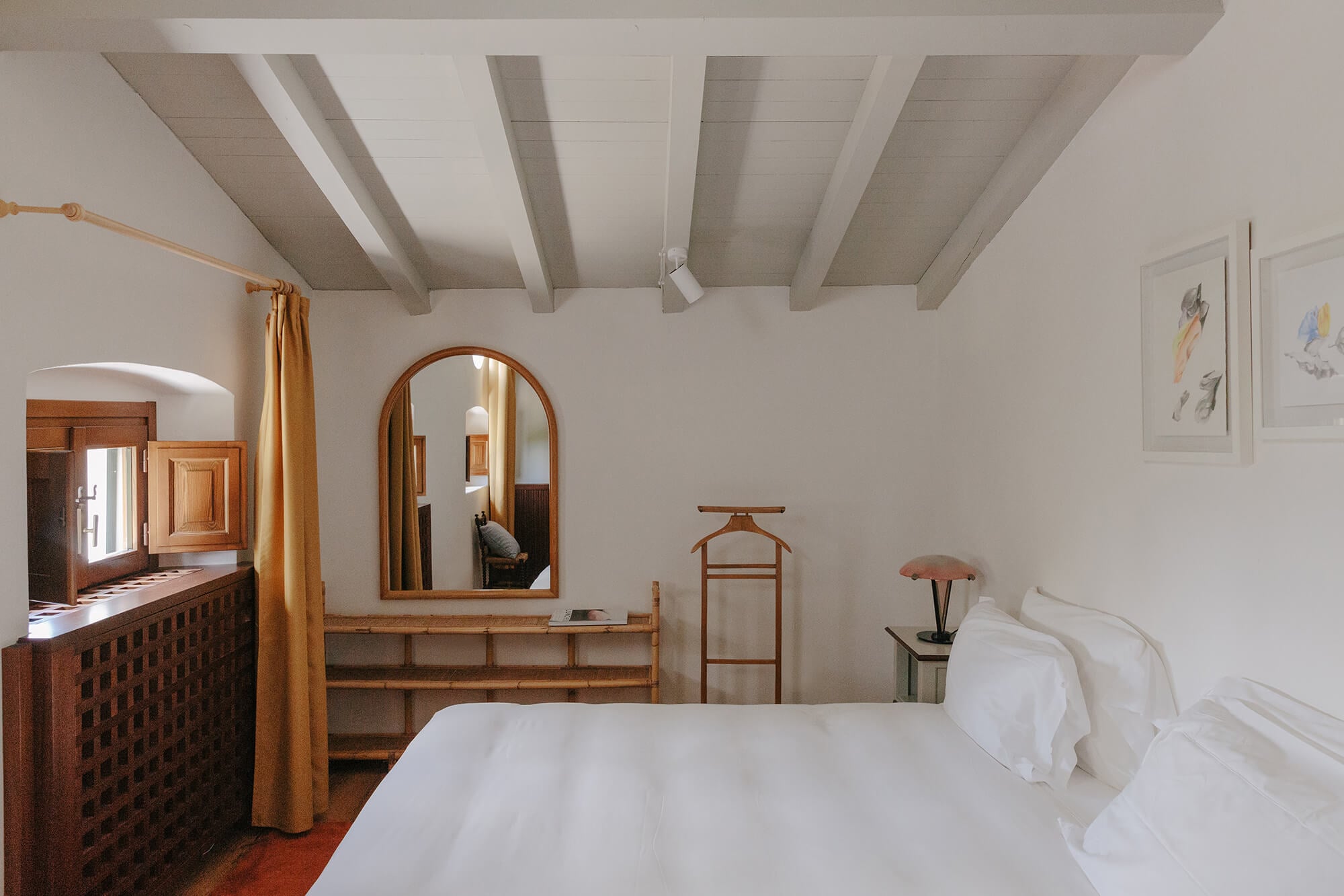 Villa Lena, Europe — Minimalist bedroom with exposed wooden beams, warm wood furniture, white linens, arched mirror, and golden curtains overlooking terracotta floors.