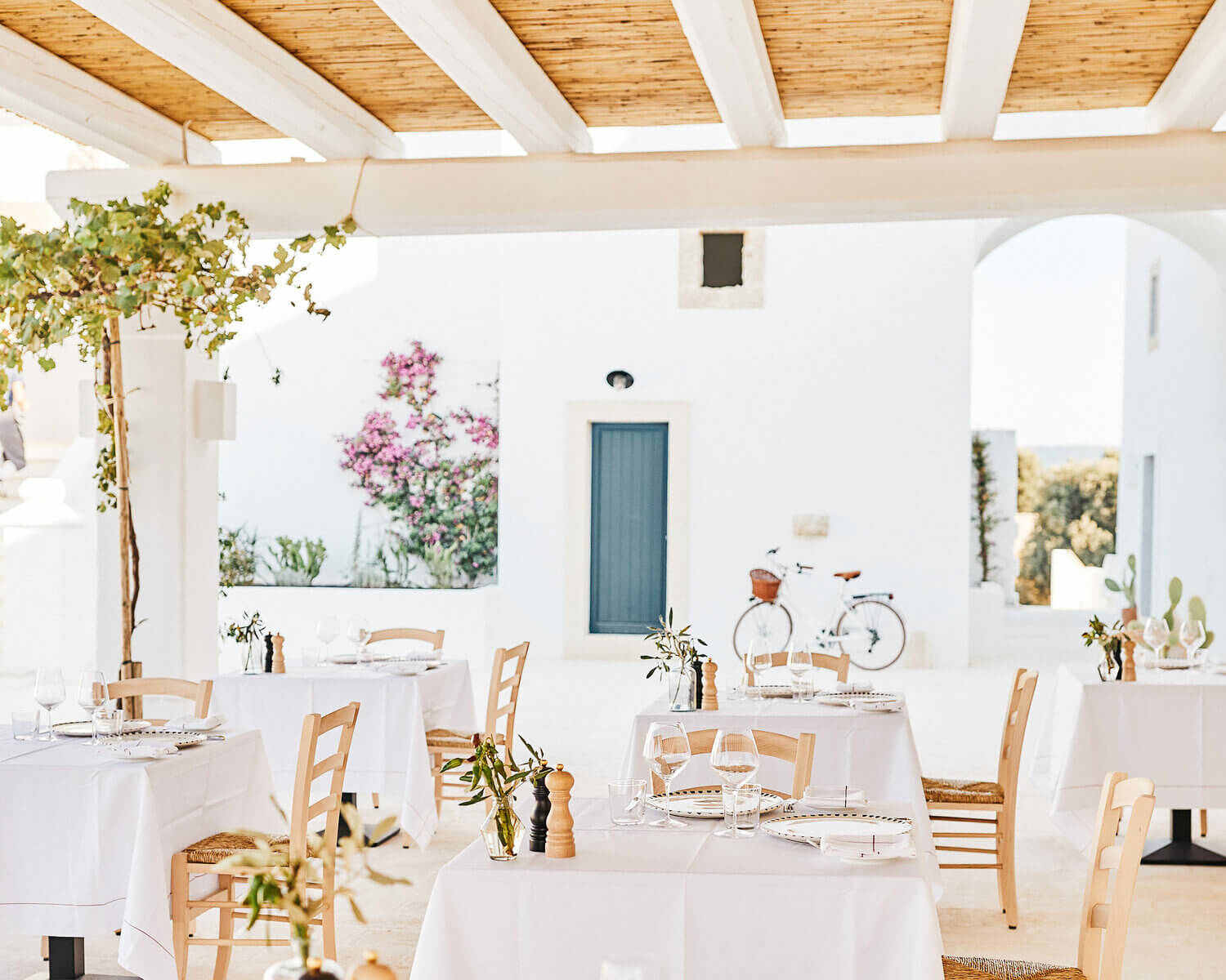 Masseria Calderisi, Europe — whitewashed Mediterranean courtyard with wooden pergola, linen-draped tables, rush-seat chairs, pink bougainvillea, and teal shuttered door.