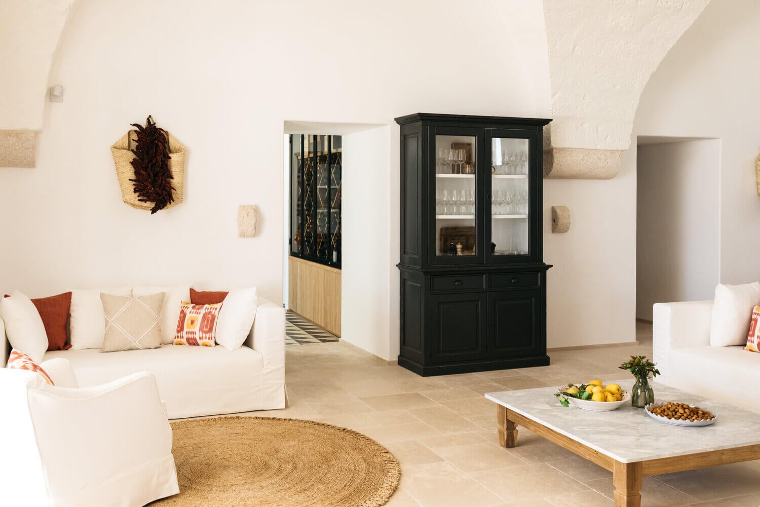 Masseria Calderisi, Europe — Bright minimalist living room with cream linen seating, black glazed cabinet, natural fiber rug, and limestone floors in Mediterranean villa.
