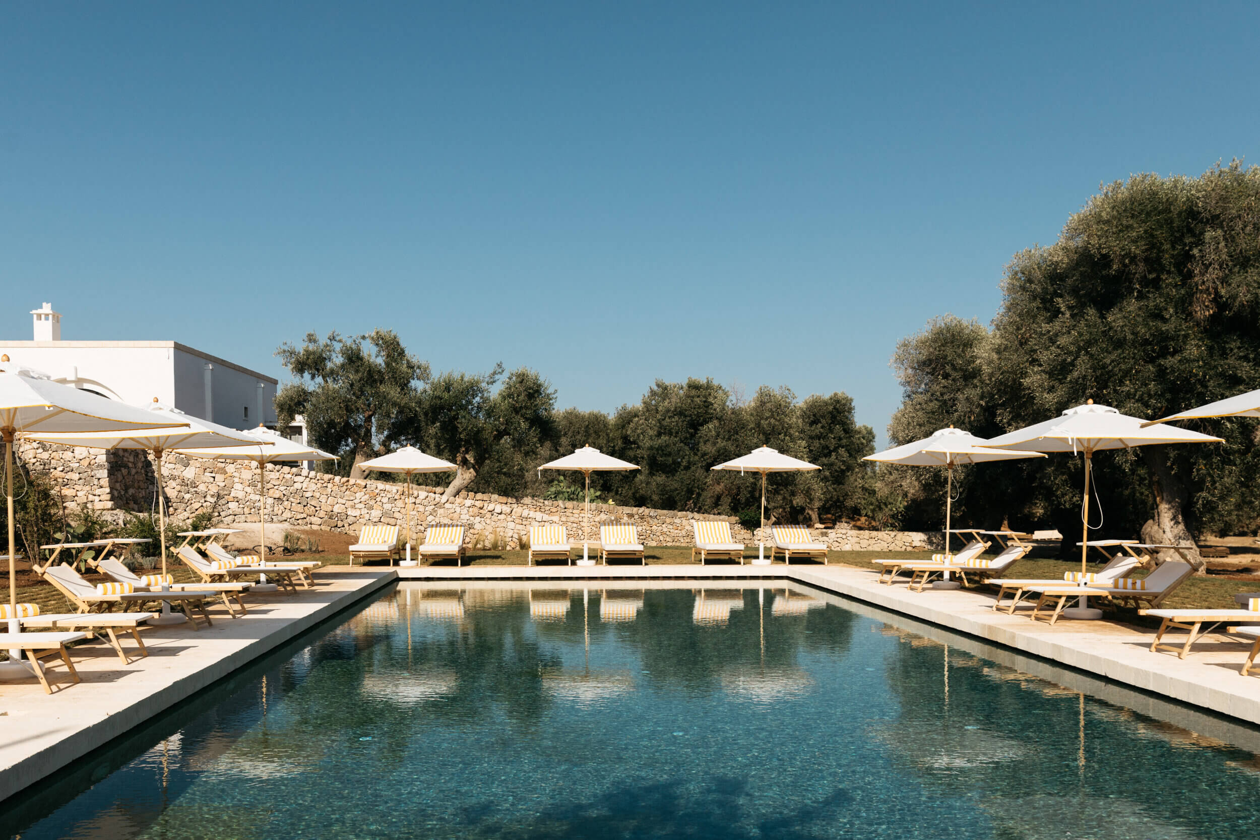 Masseria Calderisi, Europe — Linear infinity pool with natural stone terrace, wooden loungers, white umbrellas, and olive groves under clear sky.