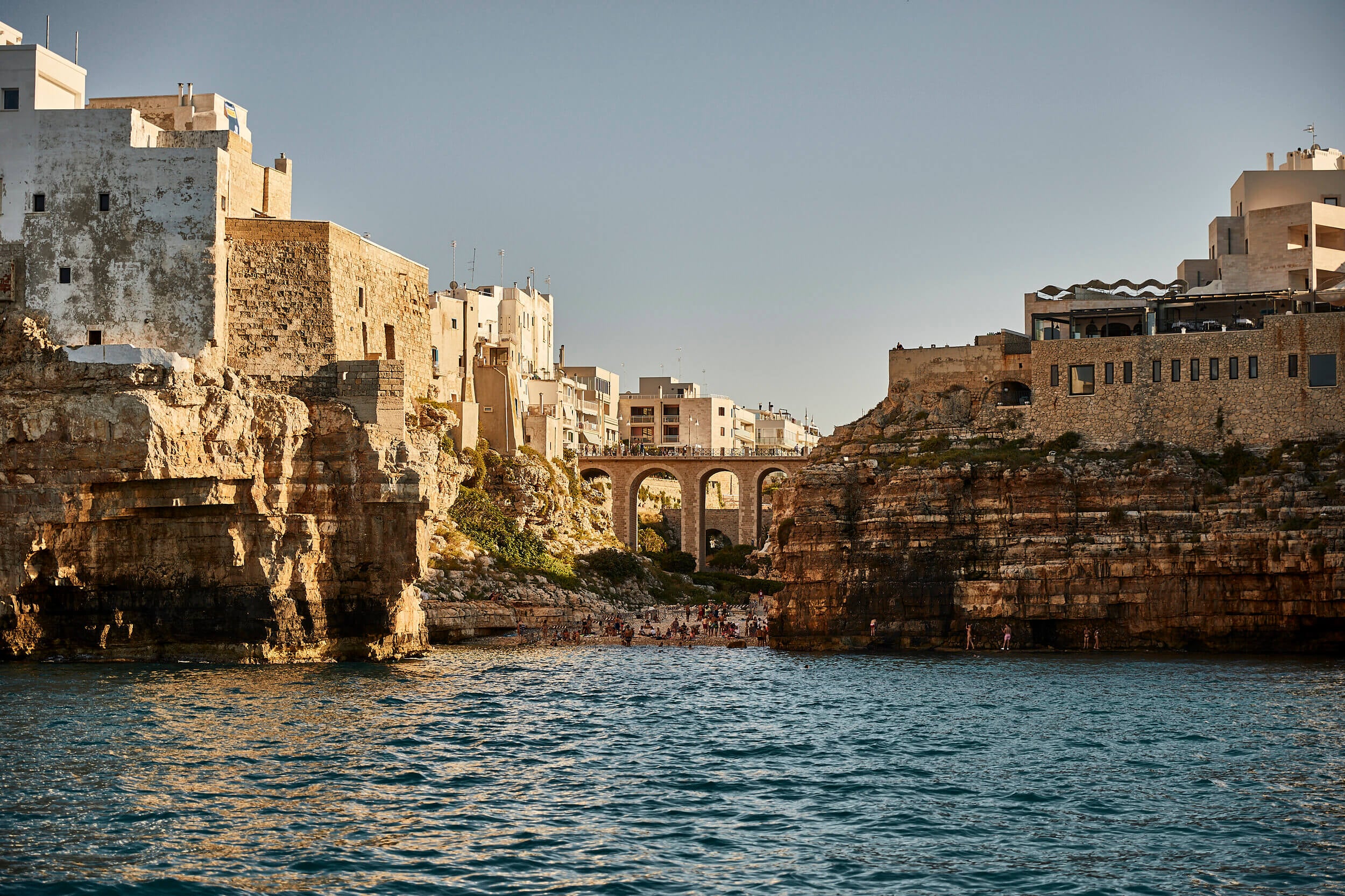 Masseria Calderisi, Europe — Limestone cliffs and arched stone bridge overlooking turquoise Mediterranean waters, historic coastal architecture bathed in golden light.