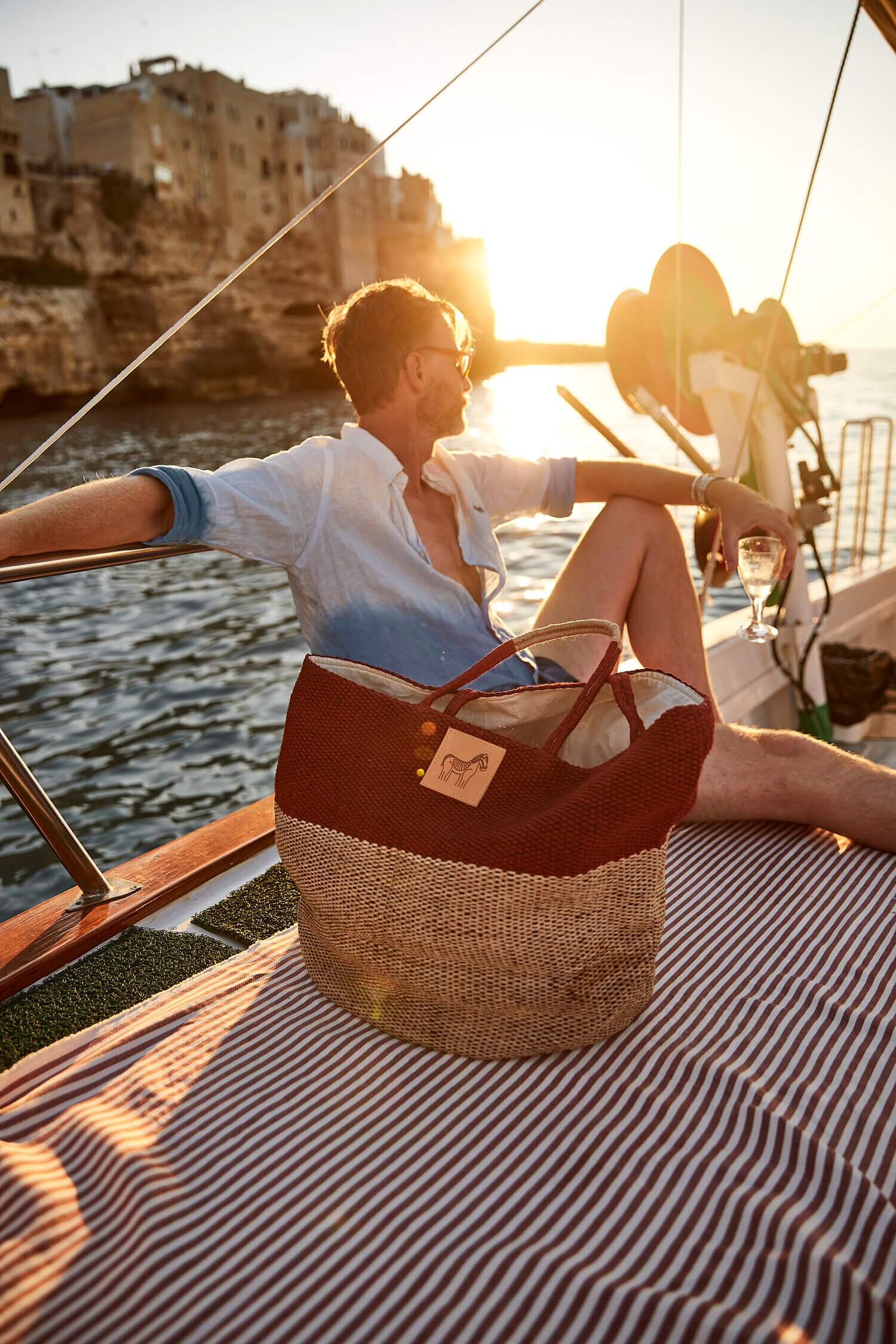 Private Boat — Man relaxing on sailing boat at sunset with coastal cliffside architecture, golden hour Mediterranean light, luxury leisure travel.