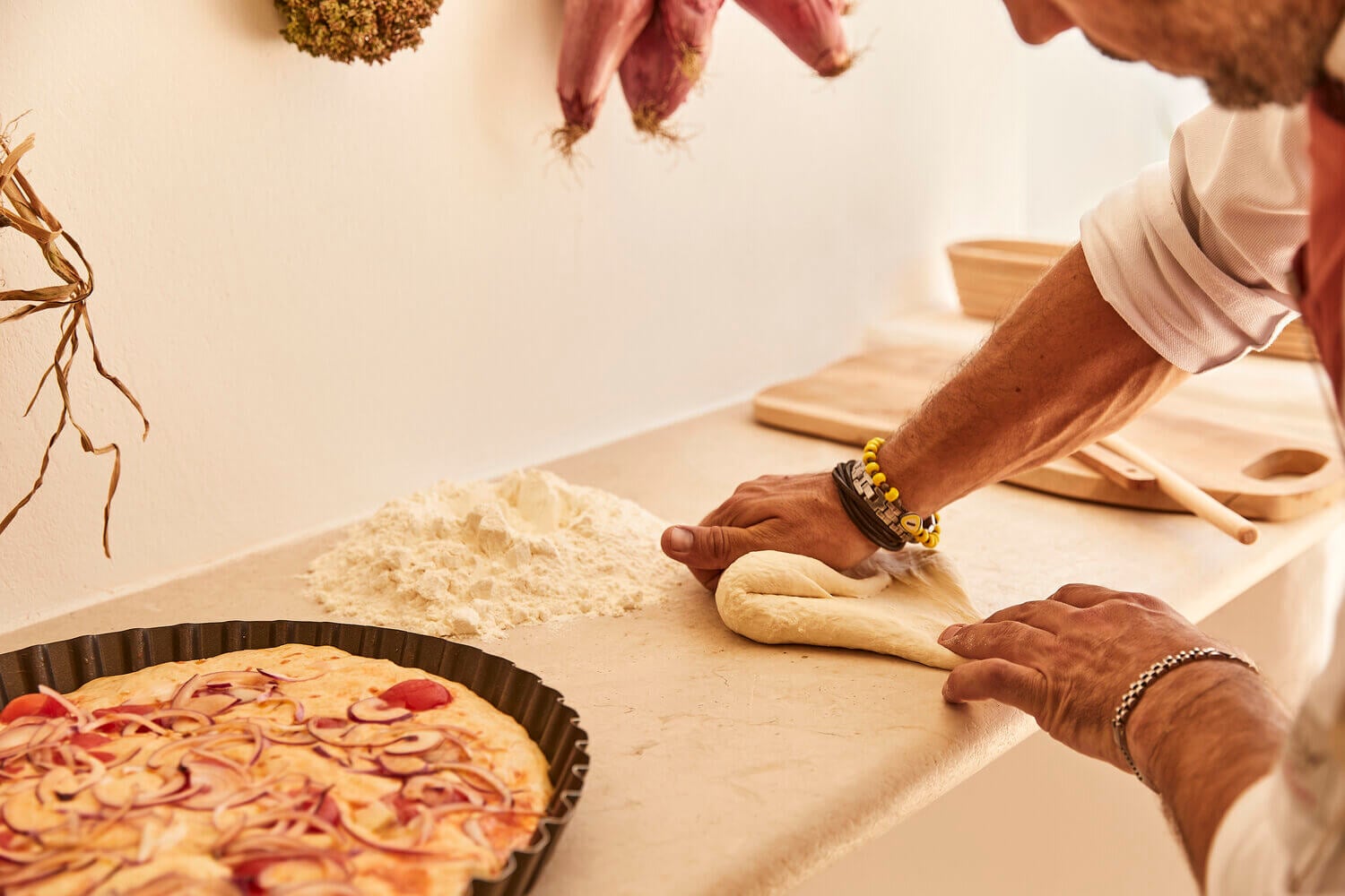 Pizza cooking class in Europe — hands shaping dough beside flour and finished focaccia, warm natural light, artisanal kitchen setup.