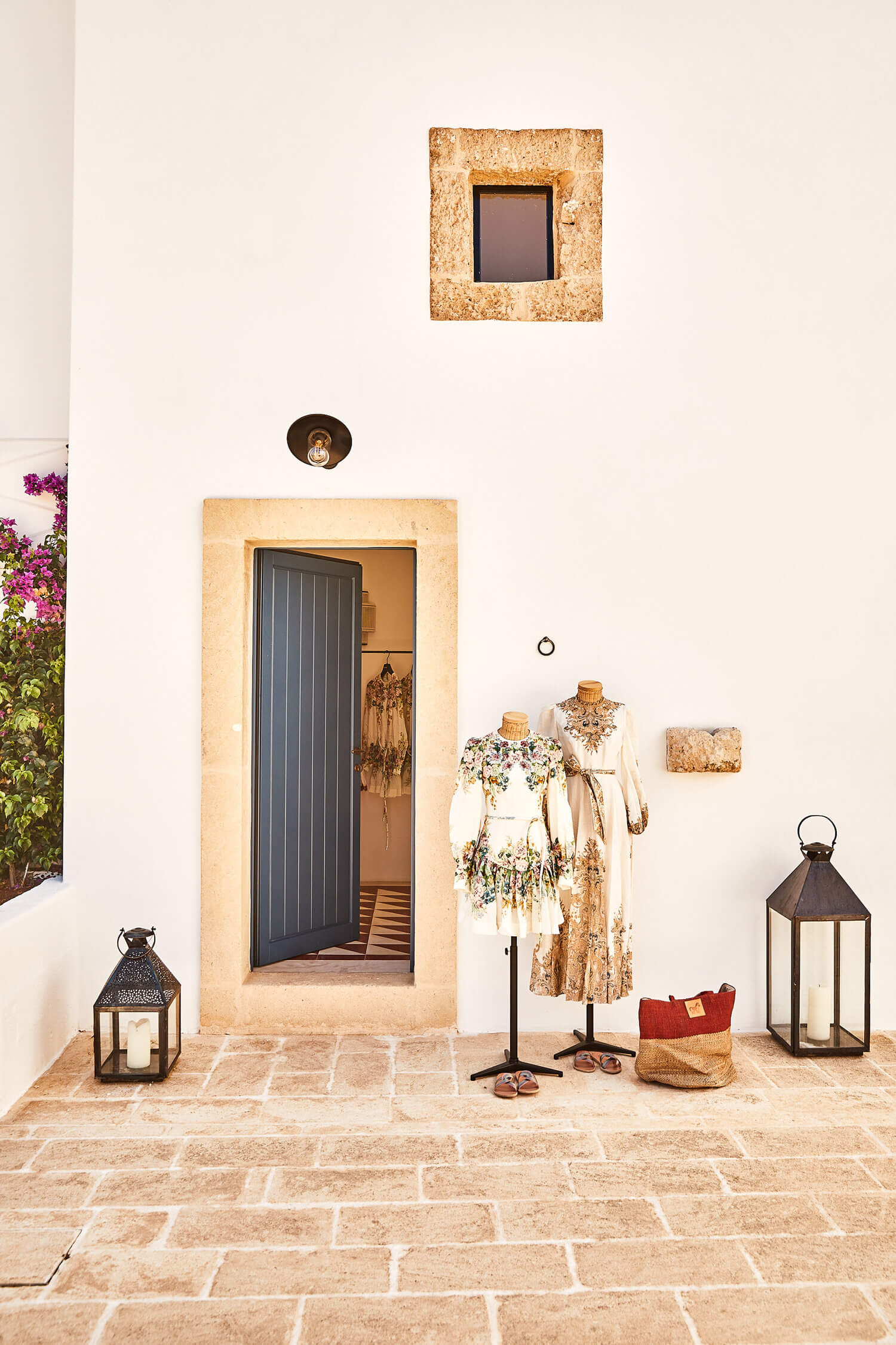 Malibu, Europe — Minimalist Mediterranean courtyard with terracotta stone paving, cream limestone archway, mannequins in floral garments, black metal lanterns, and magenta bougainvillea.