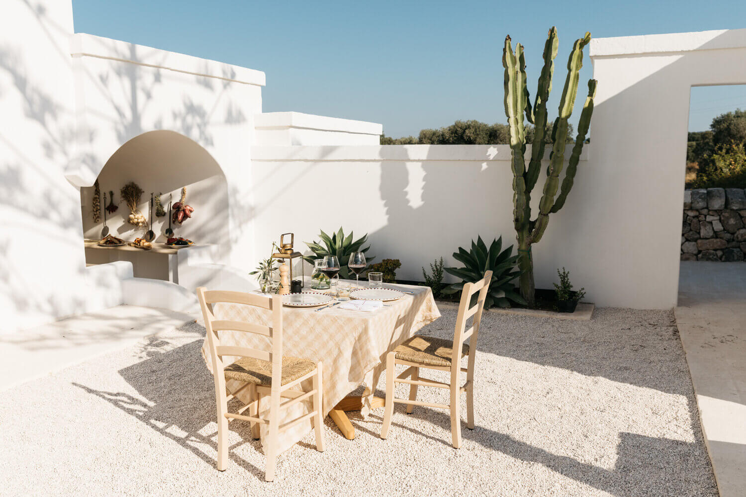 Masseria Calderisi, Europe — Whitewashed courtyard with dining table, natural wood chairs, tall cactus, and arched niche detail under Mediterranean sun.