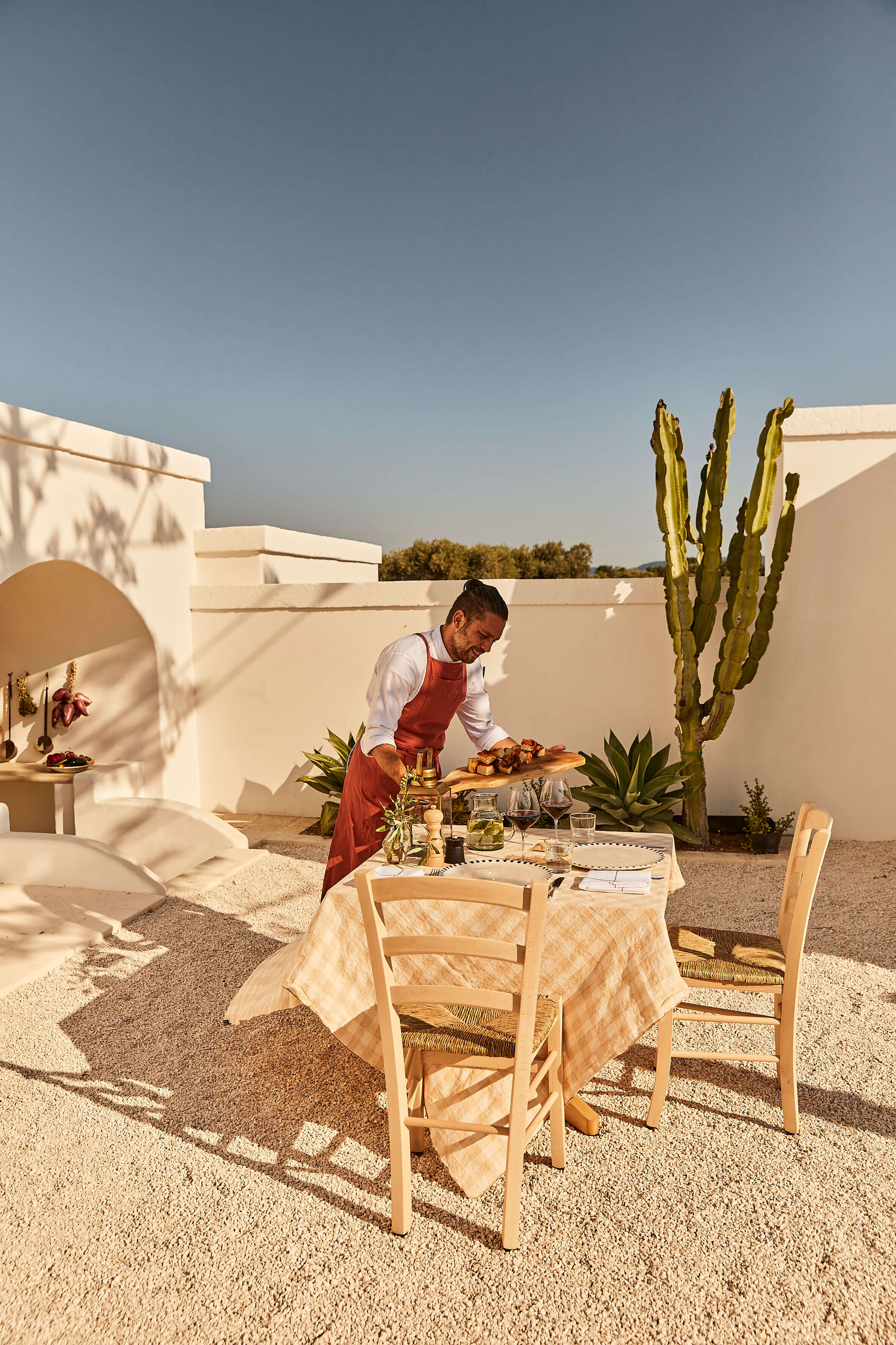 Masseria Calderisi, Europe — Chef arranging Mediterranean cuisine on linen-draped table in minimalist courtyard with whitewashed stone architecture and tall cacti.