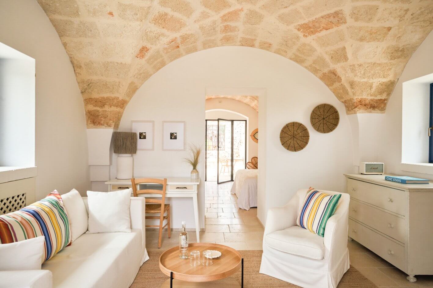 Masseria Calderisi, Europe — Whitewashed bedroom with barrel-vaulted stone ceiling, natural light, woven accents, and arched doorway opening to terracotta courtyard.