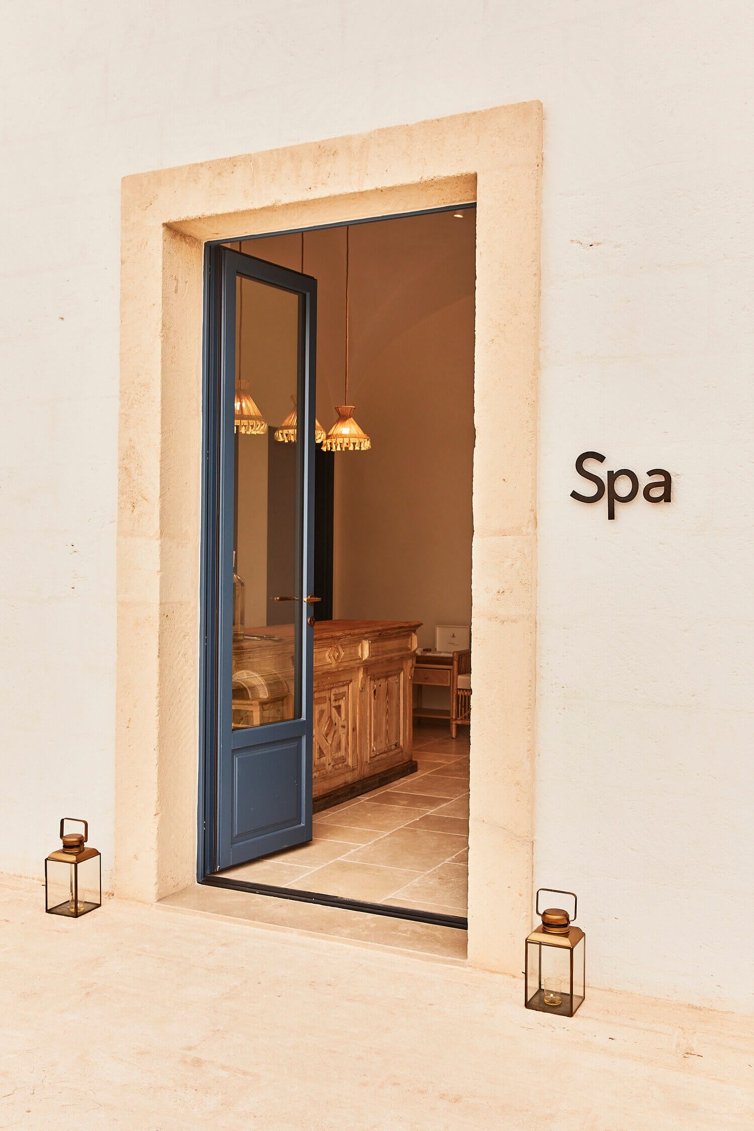 Calderisi Spa, Europe — Travertine-framed entrance with dark blue glass doors, warm terracotta interior, suspended brass pendant lights, and decorative lanterns.