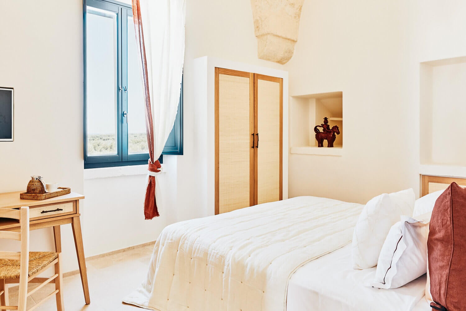 Masseria Calderisi, Europe — Minimalist bedroom with whitewashed walls, wooden wardrobes, stone archway, blue-framed window overlooking countryside, terracotta accents.