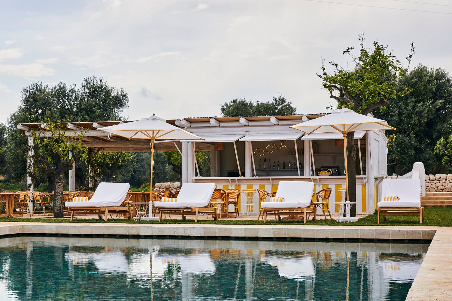 Gioia Pool Bar, Europe — Modern poolside bar pavilion with white canvas canopy, wooden loungers, striped cushions, and floating umbrellas overlooking calm water.