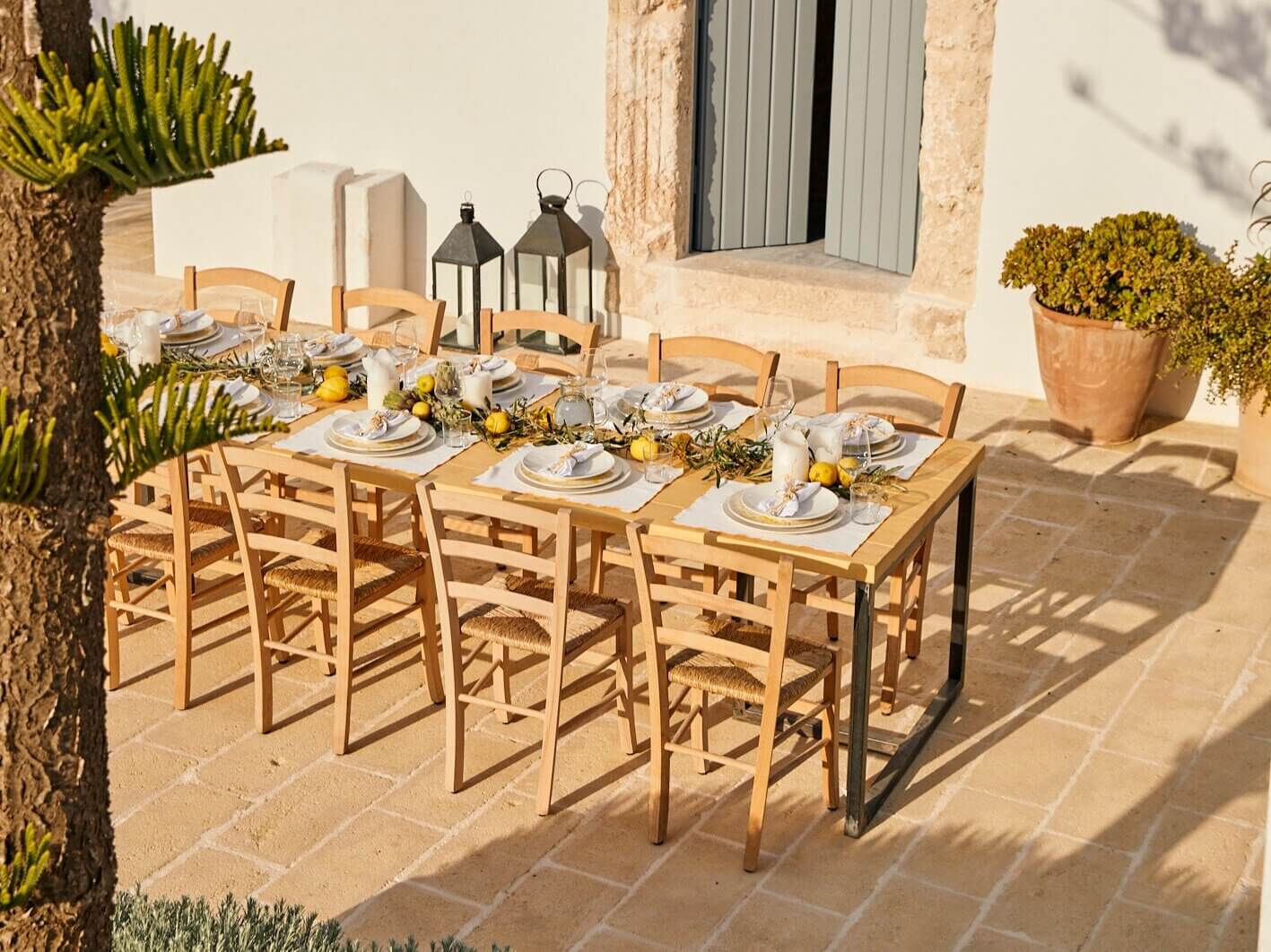 Masseria Calderisi, Europe — Terracotta courtyard dining setup with wooden chairs, white place settings, lemon centerpieces, potted succulents, and stone architecture in Mediterranean sunlight.
