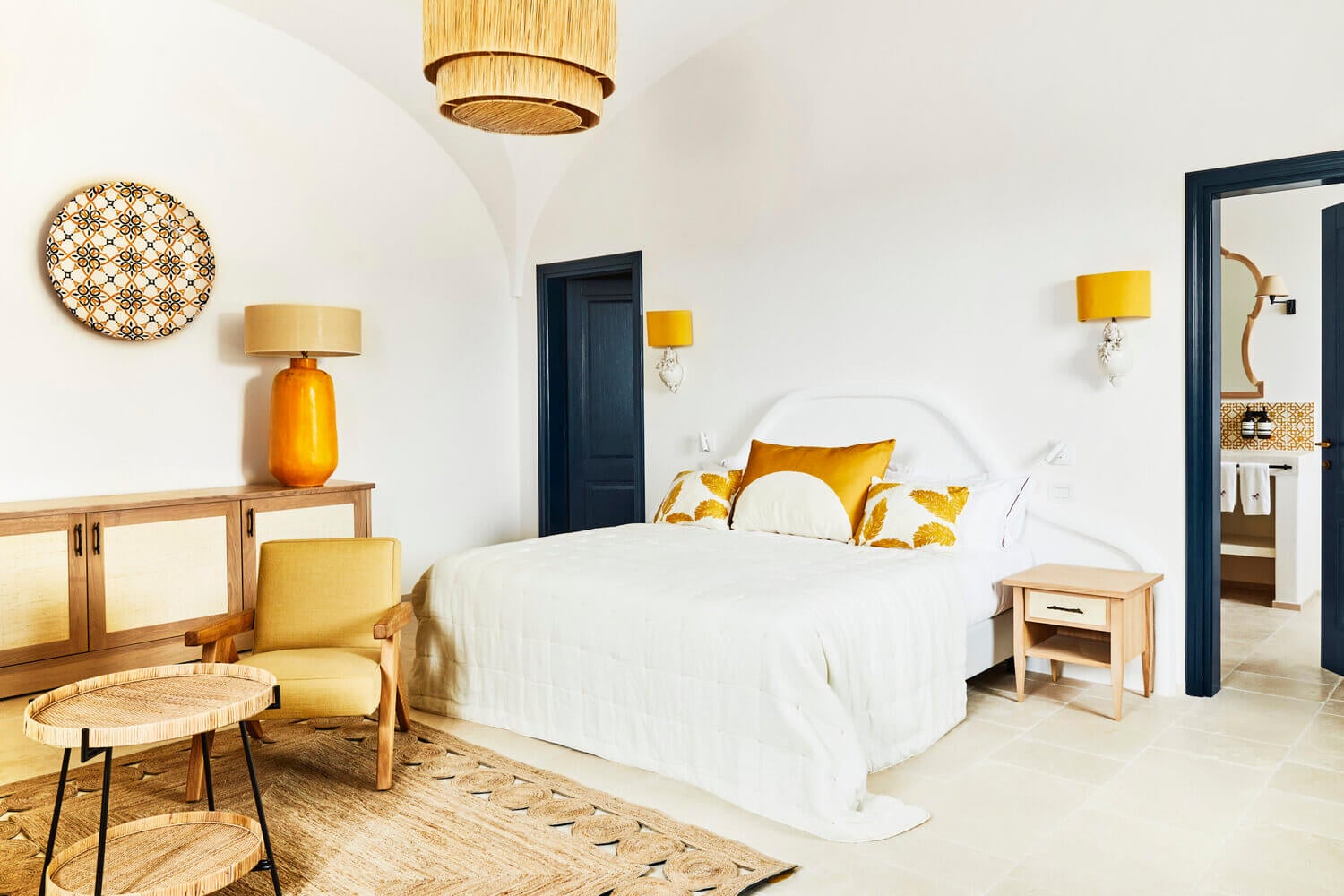 Masseria Calderisi, Europe — Sunlit bedroom with white linens, mustard accents, natural wood furniture, and dark navy doors against whitewashed walls.