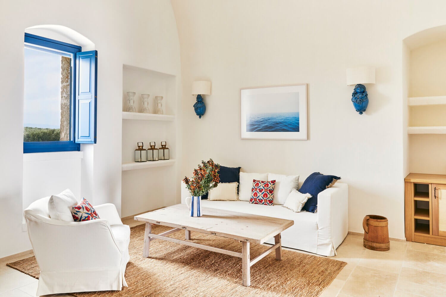 Masseria Calderisi, Europe — Whitewashed living room with blue-shuttered window, cream linen sofa, natural light, coastal artwork, woven rugs, and Mediterranean minimalist design.
