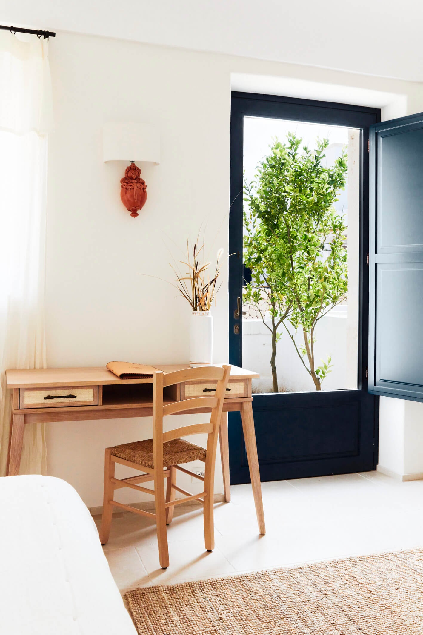 Masseria Calderisi, Europe — Minimalist bedroom with wooden desk, woven chair, terracotta wall sconce, and open doors overlooking lush green courtyard garden.