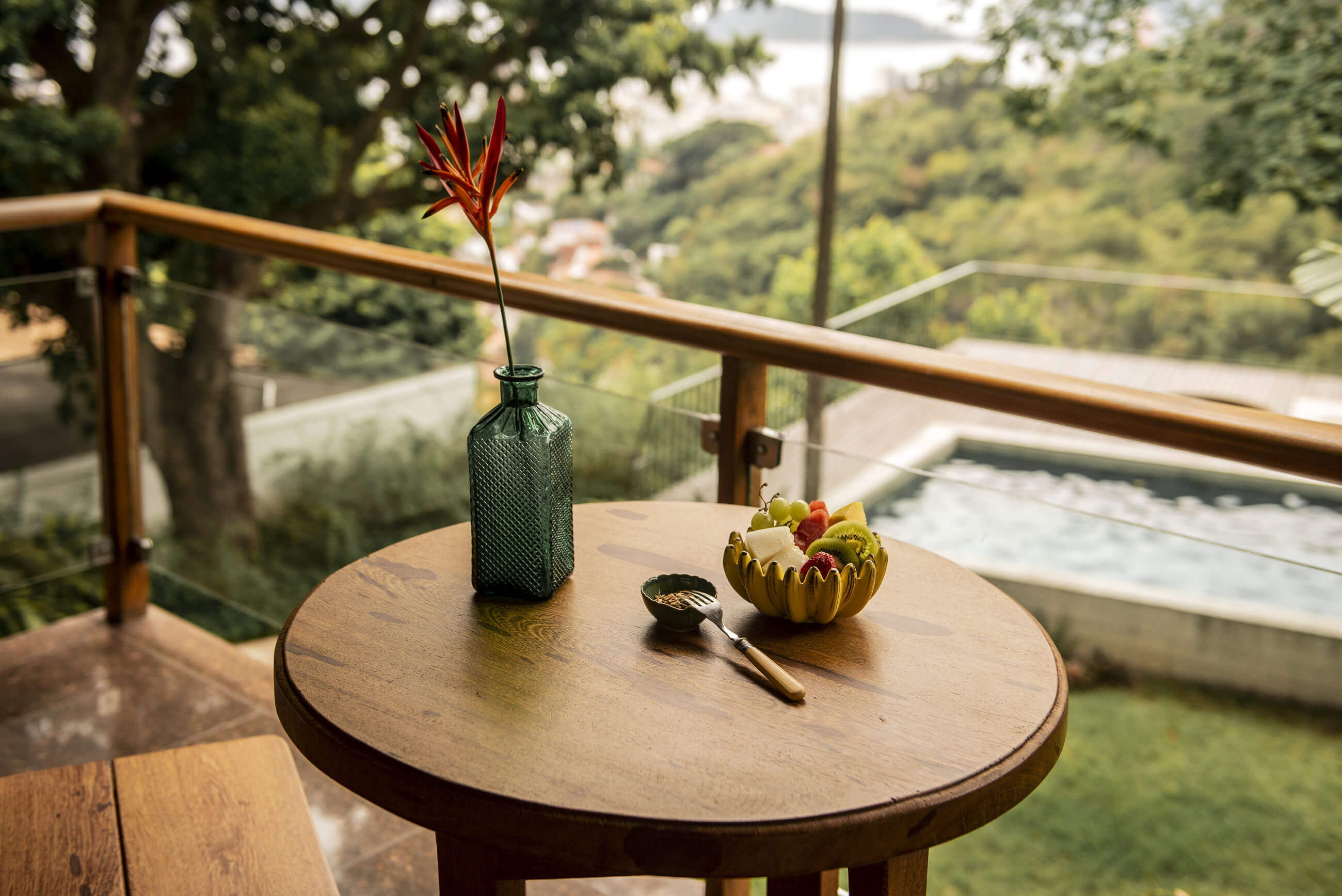 Chez Georges, South America — Round wooden table with green glass vase, red flower, and brass bowl overlooking misty valley and river from hillside terrace.