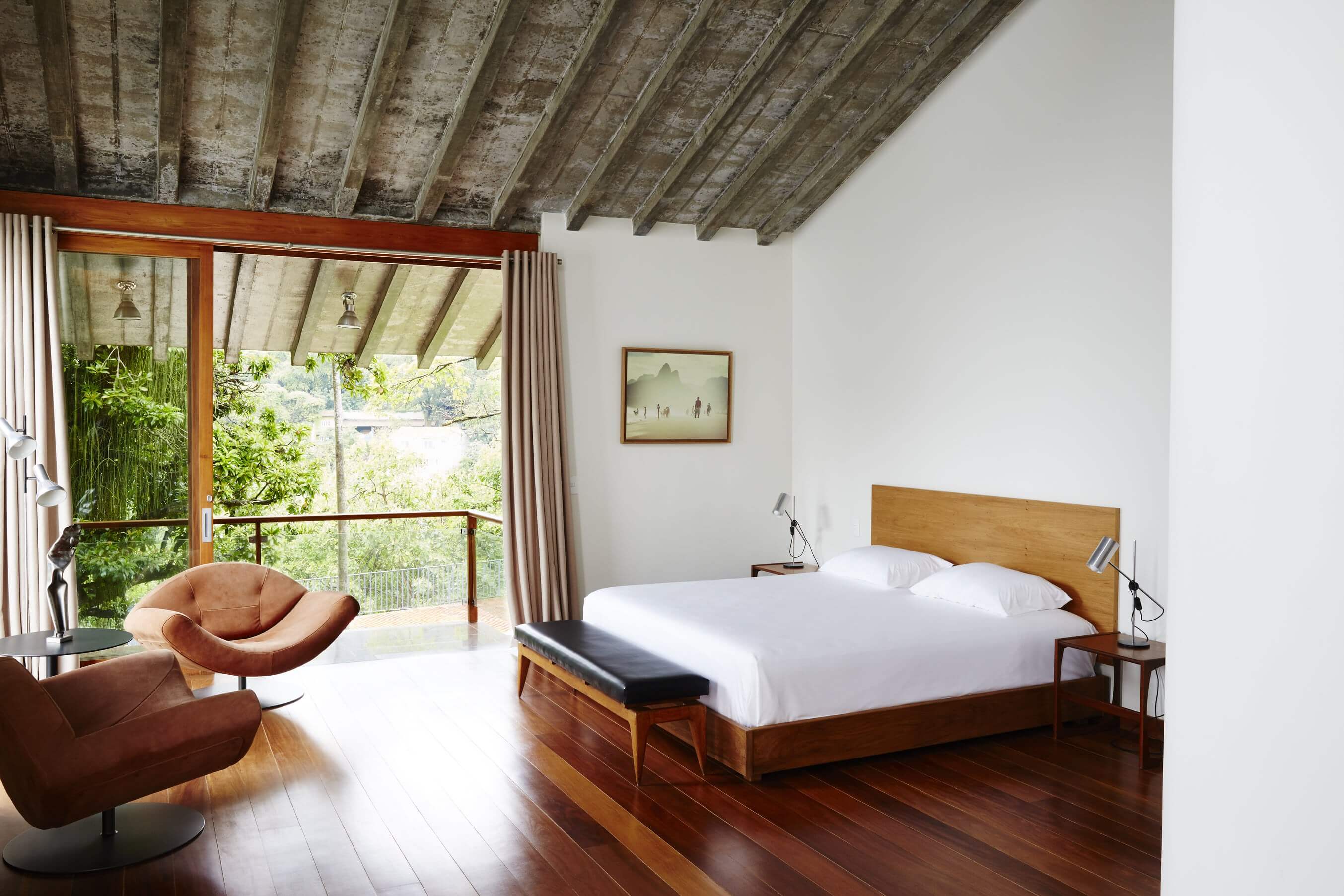 Chez Georges, South America — Modern bedroom with exposed wooden beam ceiling, floor-to-ceiling glass doors, cognac leather lounge chair, teak bed frame, and lush garden views.