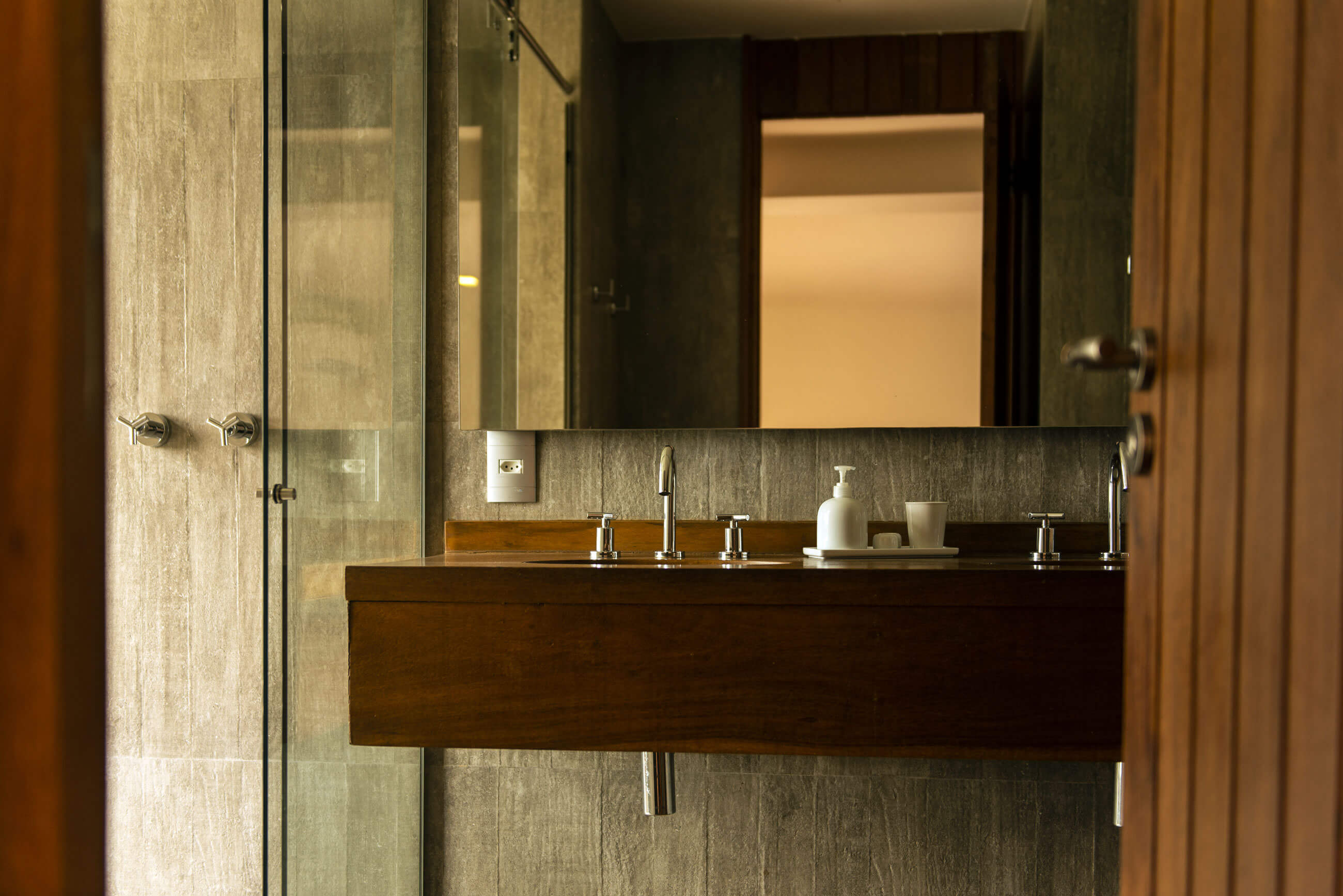 Chez Georges, South America — Modern bathroom with dark wood floating vanity, natural stone tile walls, glass shower enclosure, and brass fixtures under warm ambient lighting.