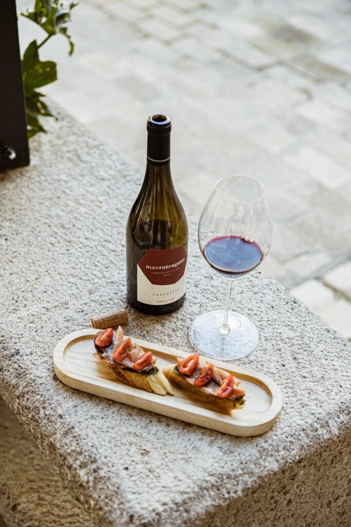 Vassaltis Vineyards, Europe — Wine bottle and glass of red wine beside charcuterie board on stone terrace overlooking white sand beach, Mediterranean coastal dining aesthetic.
