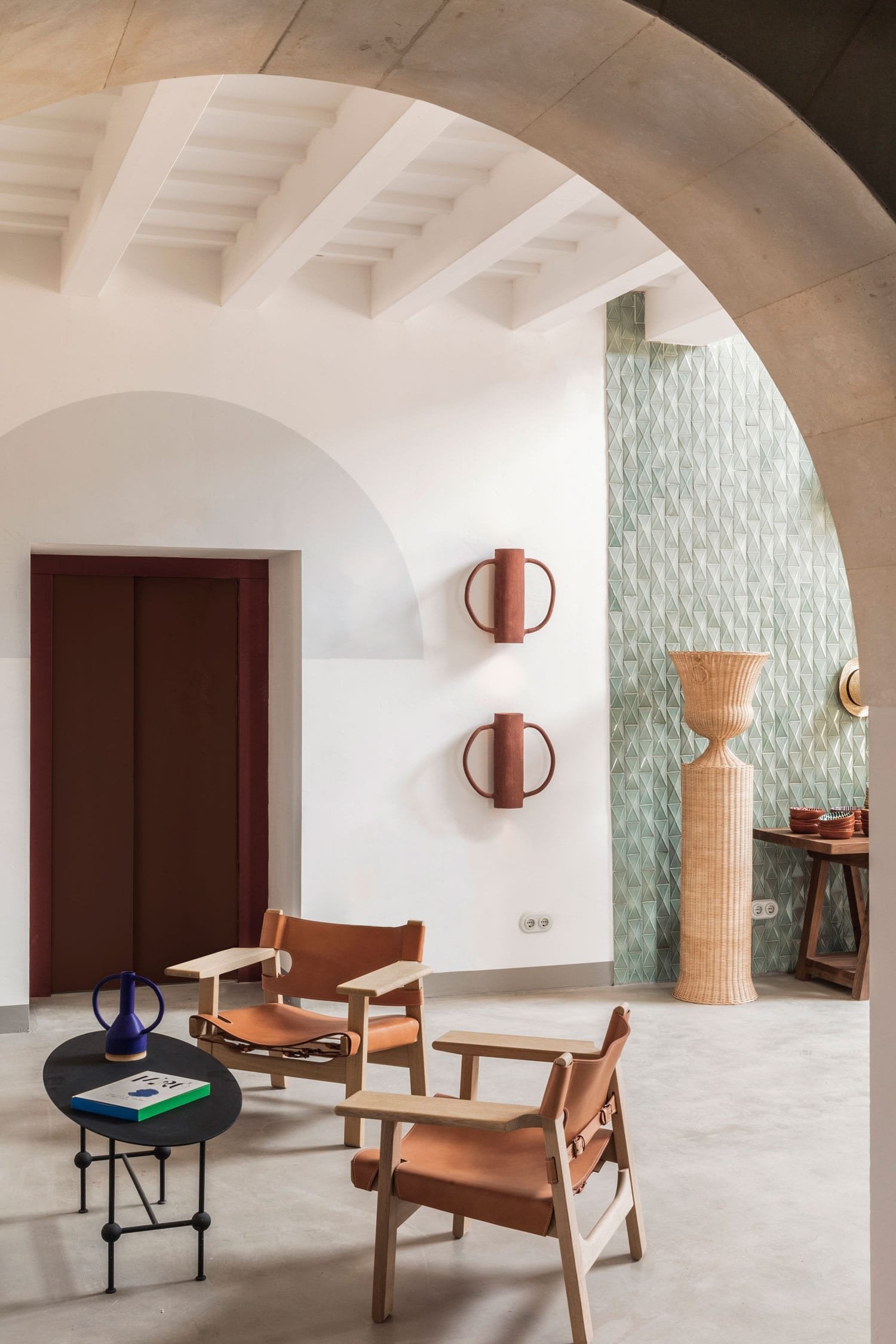 Menorca Experimental, Balearic Islands, Spain — Minimalist interior with curved white walls, terracotta leather chairs, exposed beam ceiling, patterned green wallpaper, and sculptural wooden urn.