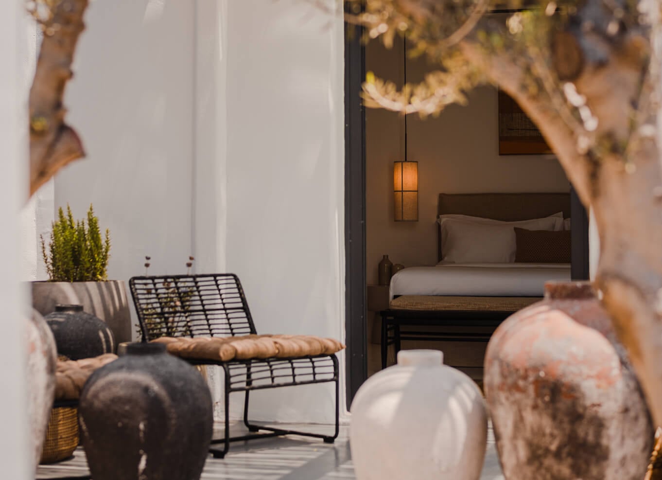 Nobu Santorini, Europe — Whitewashed courtyard with black woven chair, terracotta vessels, and glimpse of minimalist bedroom with pendant light through open doors.