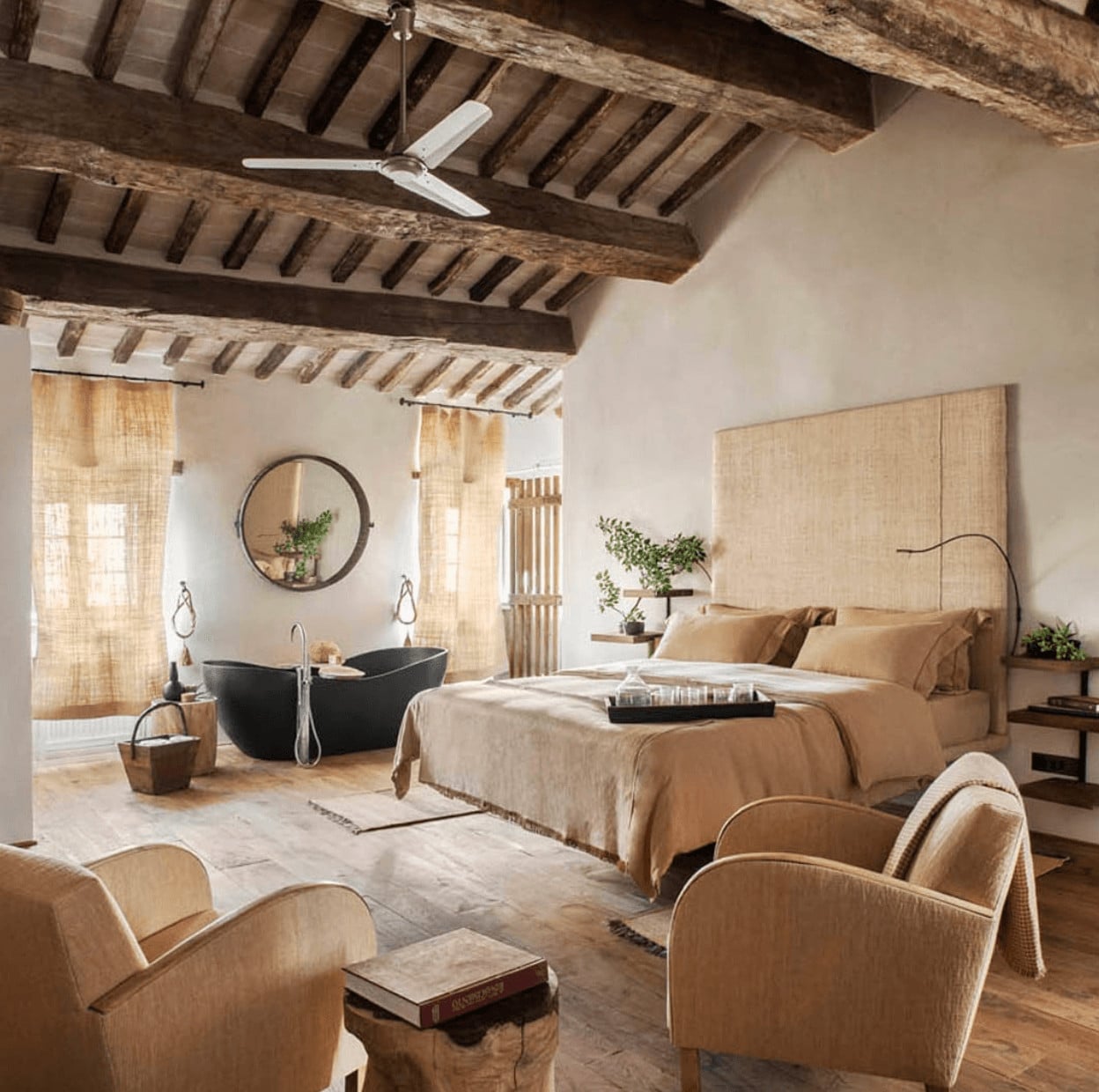 Monteverdi, Europe — Rustic bedroom suite with exposed wooden beams, neutral linens, black soaking tub, natural light through woven shades, and curated furnishings.