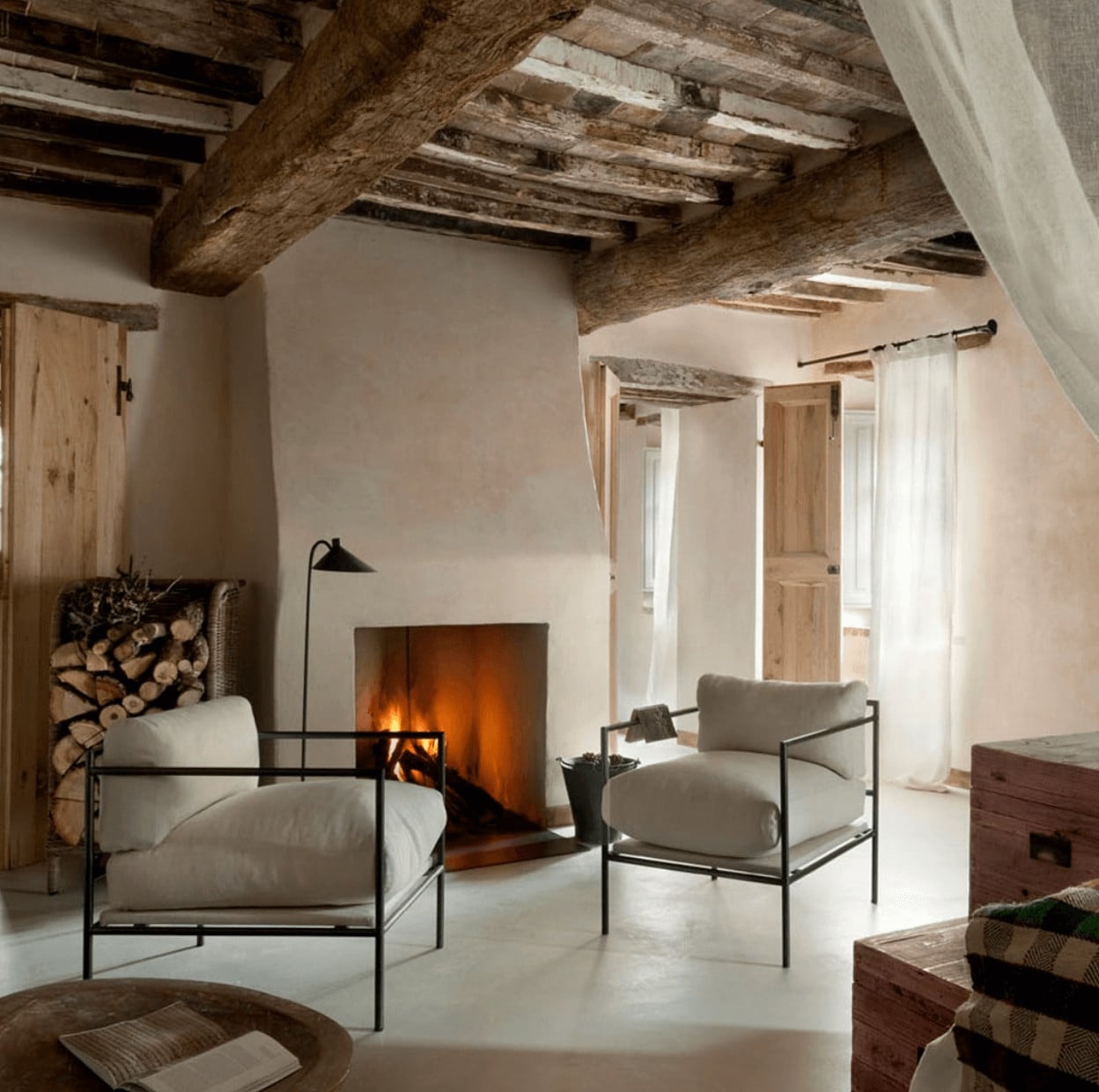 Monteverdi, Europe — Rustic stone beams frame fireplace seating area with cream linen chairs, wooden doors, and warm ambient lighting in minimalist interior.