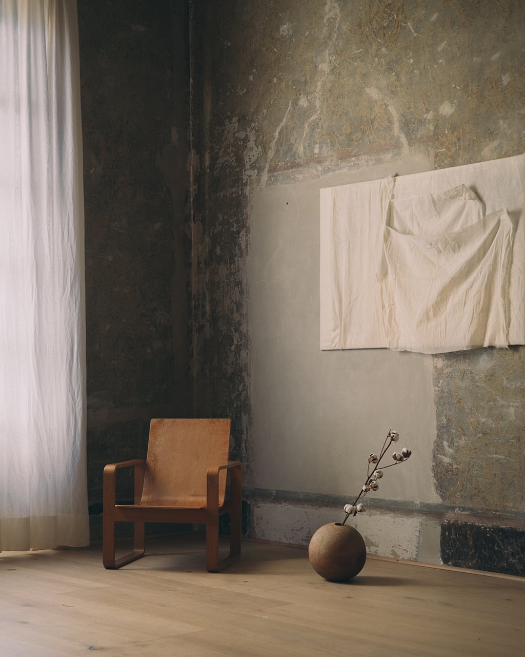 Numeroventi, Europe — Minimalist interior with cognac leather chair, dried botanical in earthenware vessel, cream linens on weathered plaster wall, natural light.