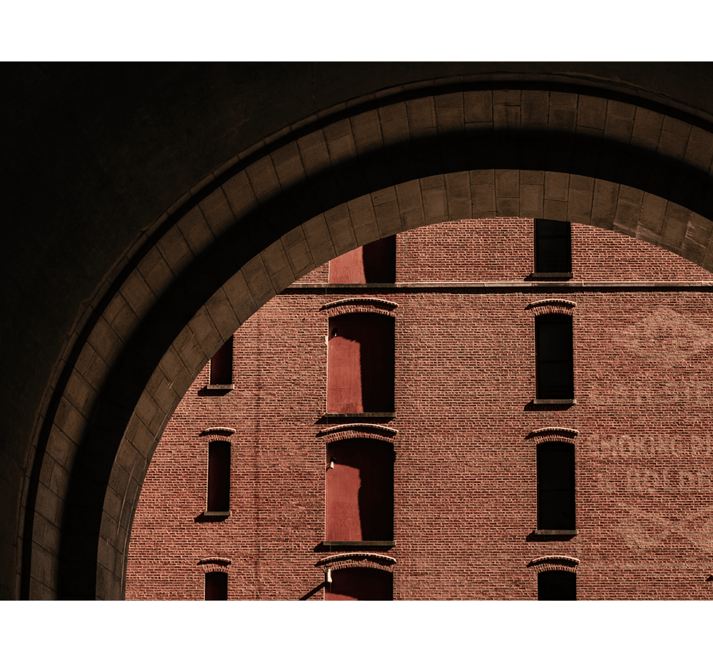 168 Plymouth, North America — Arched brick passageway framing red-brick facade with symmetrical windows and warm afternoon light casting geometric shadows.