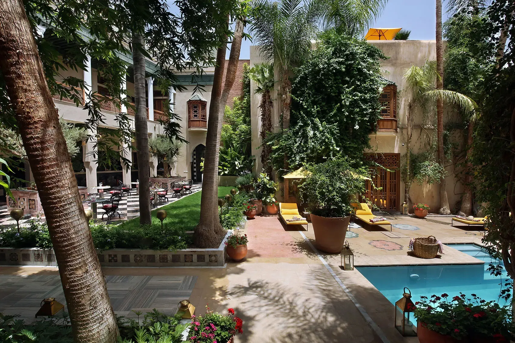 El Fenn, Marrakech, Morocco — Courtyard with palm trees, blue pool, ivy-covered walls, yellow loungers, traditional carved doors, and checkered floor in afternoon light.