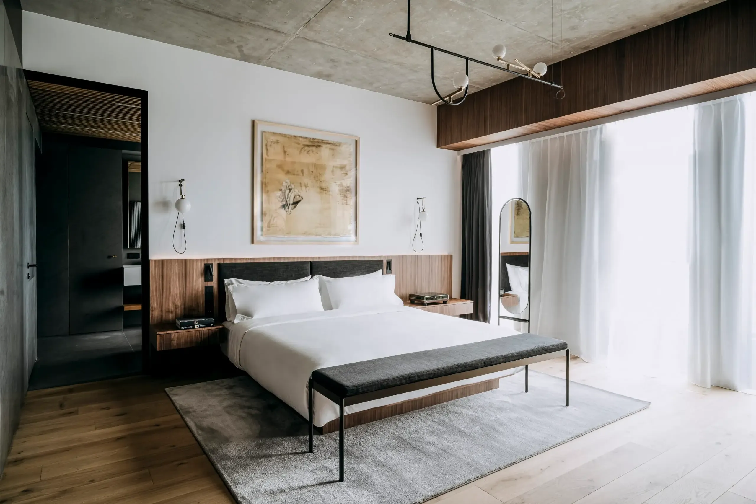 Nobu Warsaw, Poland — Modern luxury bedroom with walnut headboard, concrete ceiling, pendant lighting, and floor-to-ceiling windows with sheer curtains.