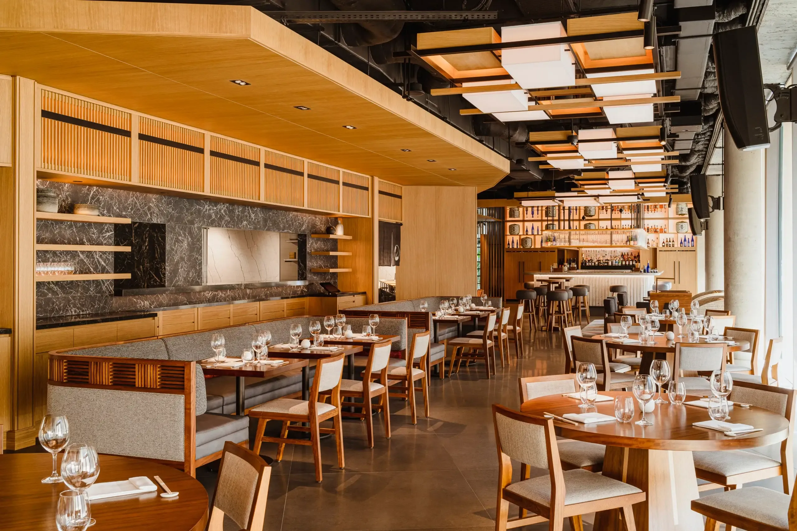 Nobu Warsaw, Poland — Warm wood ceiling with linear pendant lights above dining room featuring marble accent wall, leather banquettes, and mid-century modern chairs.