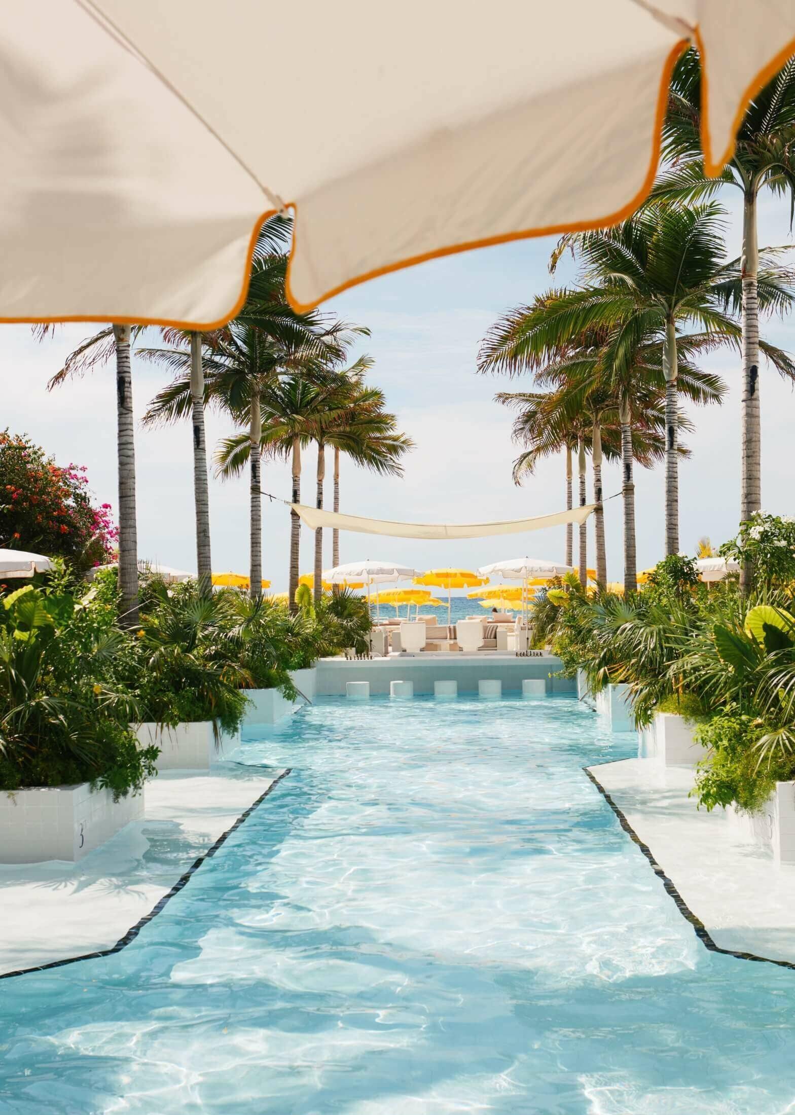 Palm Heights, North America — Illuminated swimming channel flanked by palms and orange shade sails, leading to beach loungers and ocean view.