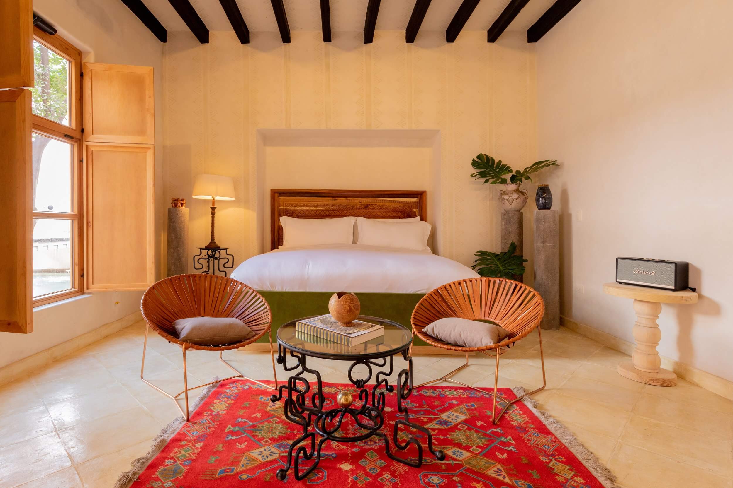 La Valise San Miguel, Mexico — Minimalist bedroom with wooden beam ceiling, terracotta chairs, red geometric rug, and natural light from arched doorway.