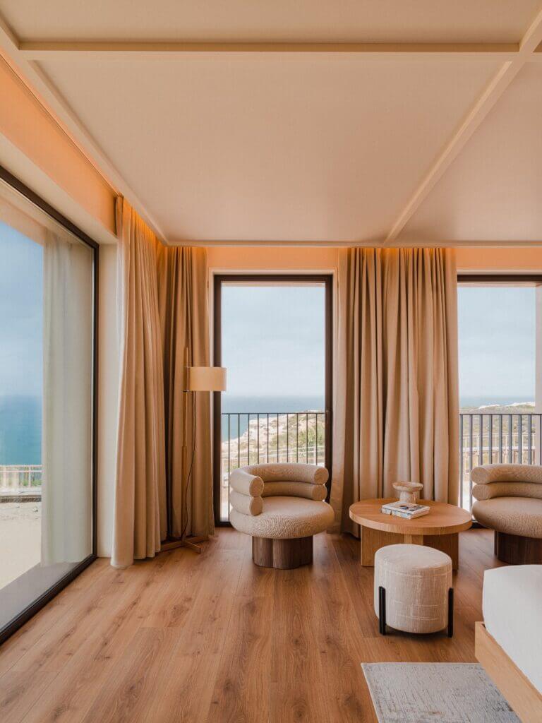 Aethos Ericeira, Portugal — Coastal suite with golden curtains, curved seating, wooden flooring, and ocean views through glass doors.