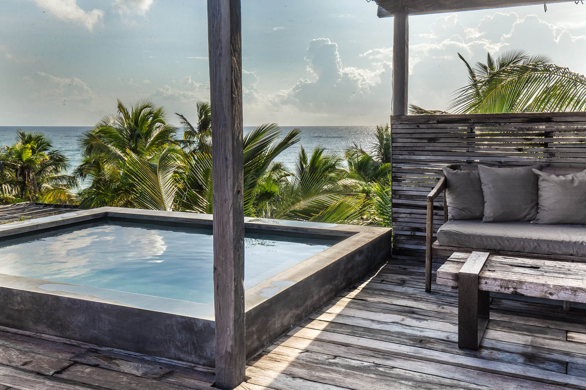 Be Tulum, Mexico — Oceanfront deck with infinity pool, wooden pergola, palm trees, and gray upholstered daybed overlooking Caribbean Sea.