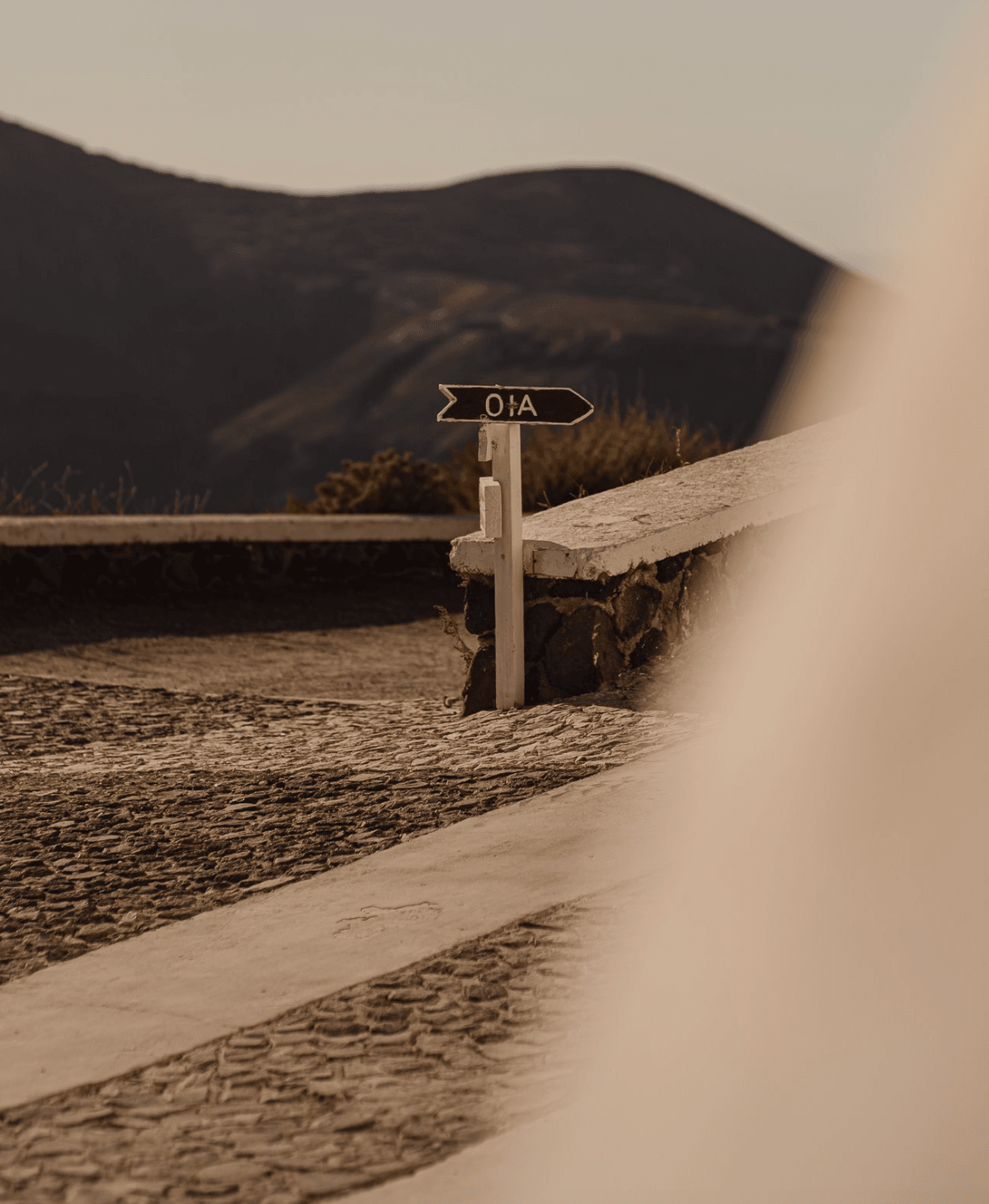 Oia – Thira Hiking Trail — Oia hiking trail directional wooden sign on sandy path with dark volcanic mountain backdrop and coastal vegetation, Santorini Greece.