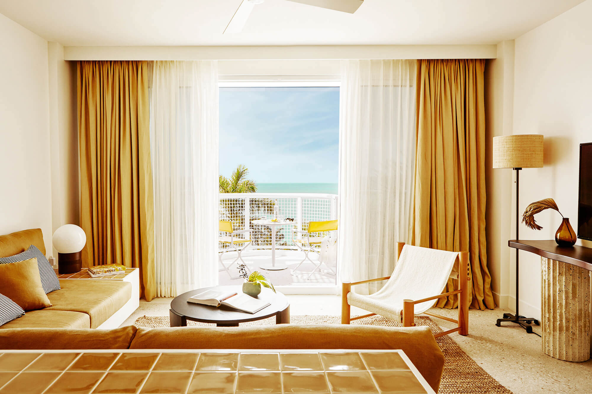 Palm Heights, North America — Oceanfront suite with gold curtains, mid-century lounge chair, and turquoise sea view from balcony.