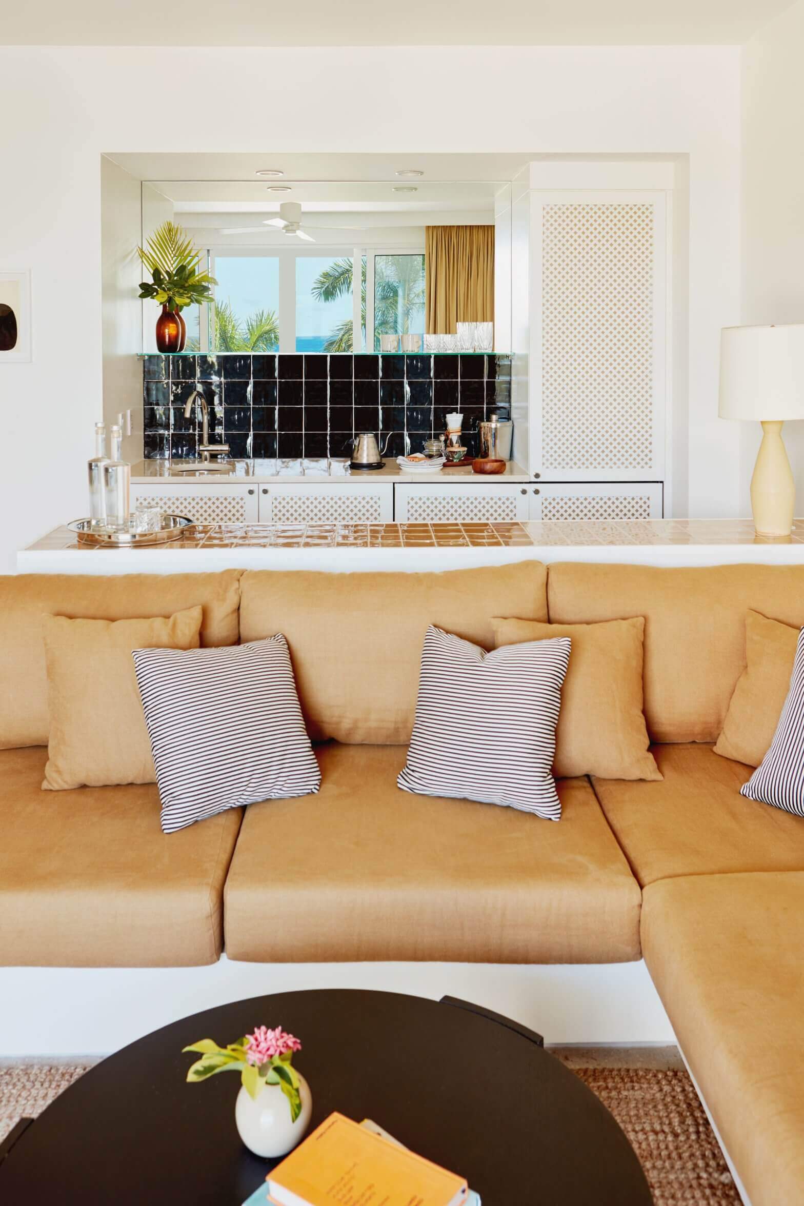 Palm Heights, North America — Coastal living room with tan leather sectional, striped pillows, black marble wet bar, ocean views, and golden curtains.