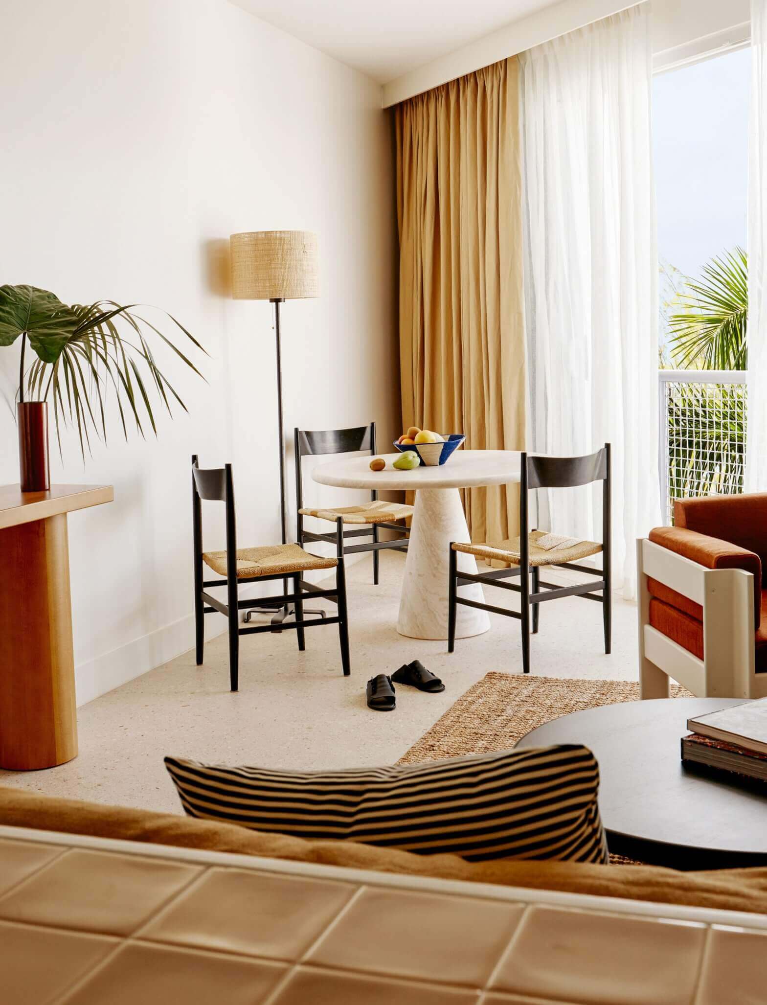 Palm Heights, North America — Modernist hotel room with round travertine dining table, woven rush chairs, warm terrazzo flooring, natural light, potted palms, and gold curtains.