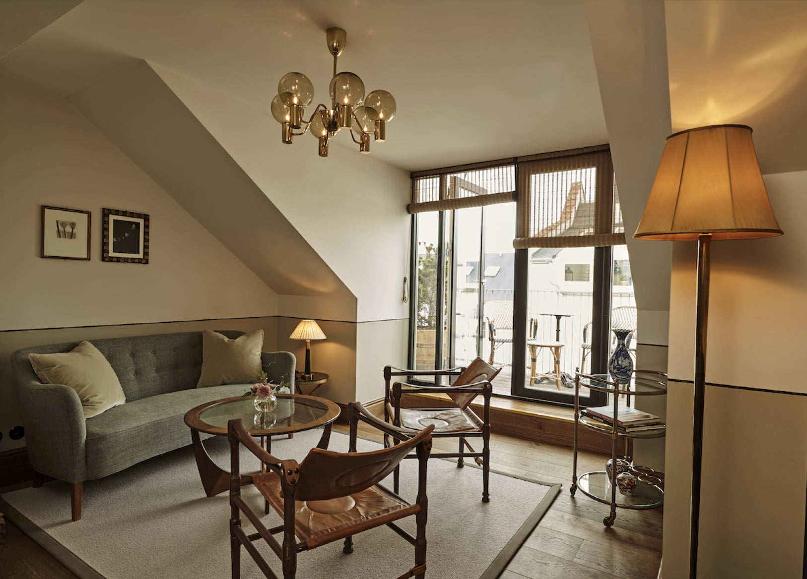 Hotel Sanders, Europe — Sloped-ceiling suite with curved green sofa, mid-century wooden chairs, round side table, brass pendant chandelier, floor lamp with amber shade, and glazed doors opening to rooftop terrace with city views.