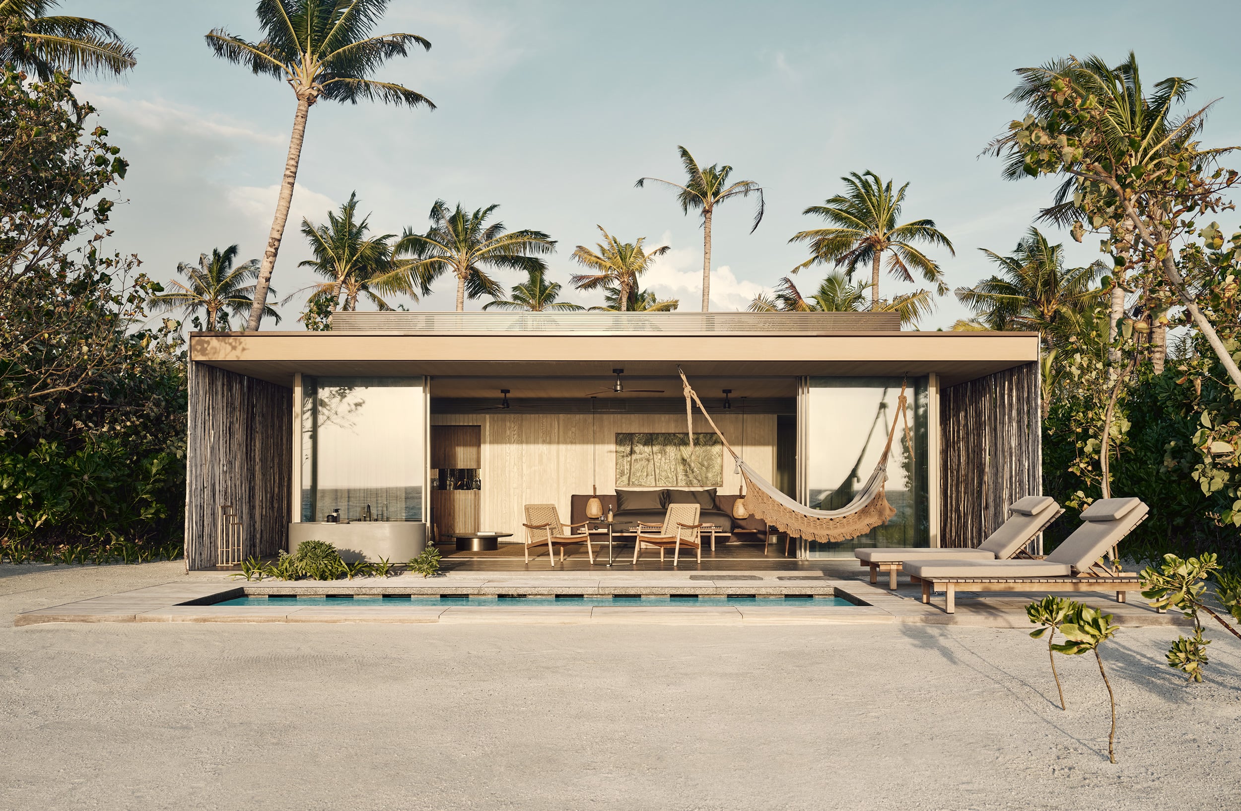 Patina Maldives, Asia — Modern beachfront villa with weathered timber facade, expansive glass doors, hanging hammock, lounge chairs, and infinity pool overlooking tropical palms.