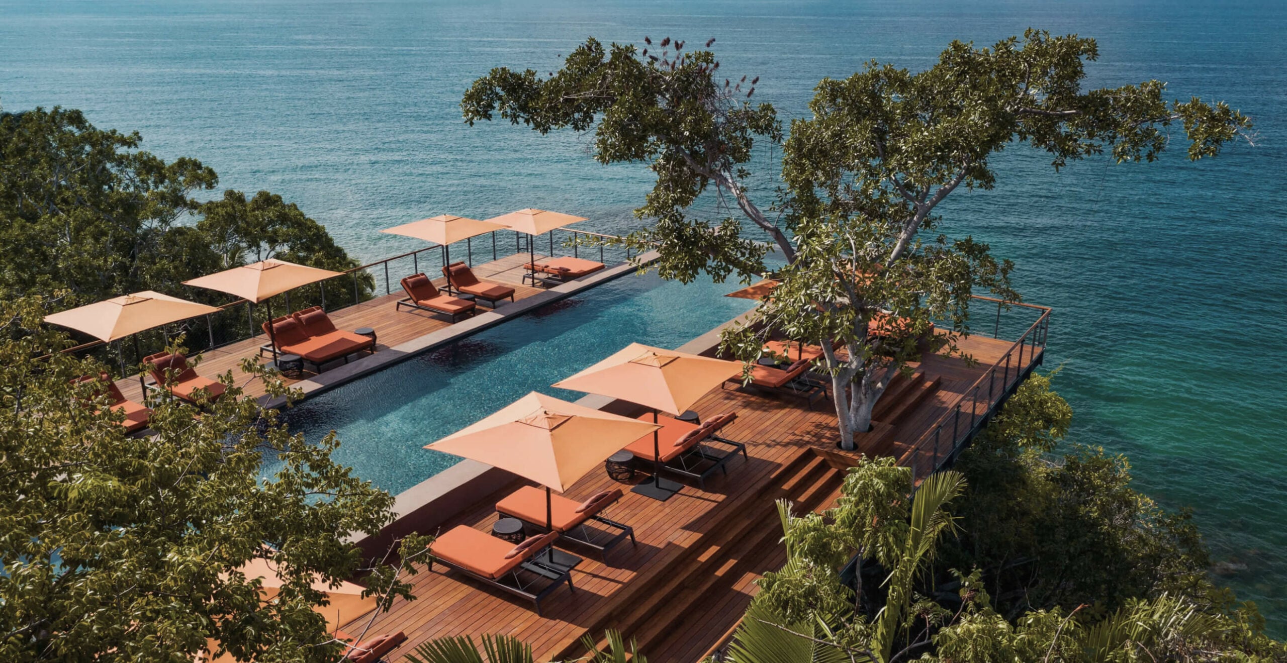 One&Only Mandarina, Mexico — Terraced wooden deck with orange loungers and umbrellas overlooking infinity pool and turquoise ocean, framed by native trees.