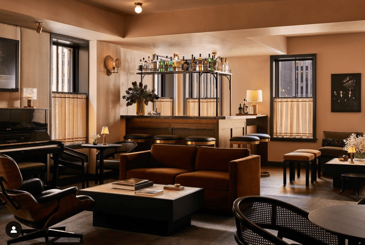 Pebble Bar, North America — Mid-century lounge with cognac leather sofa, dark wood bar, brass shelving, soft ambient lighting, and cream curtains framing windows.
