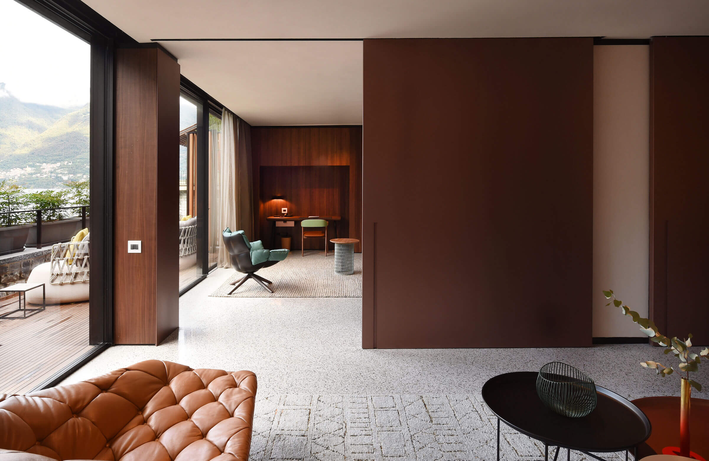 il Sereno Lago di Como, Italy — Modern luxury suite with warm wood paneling, cognac leather seating, and floor-to-ceiling windows overlooking alpine landscape and lake views.