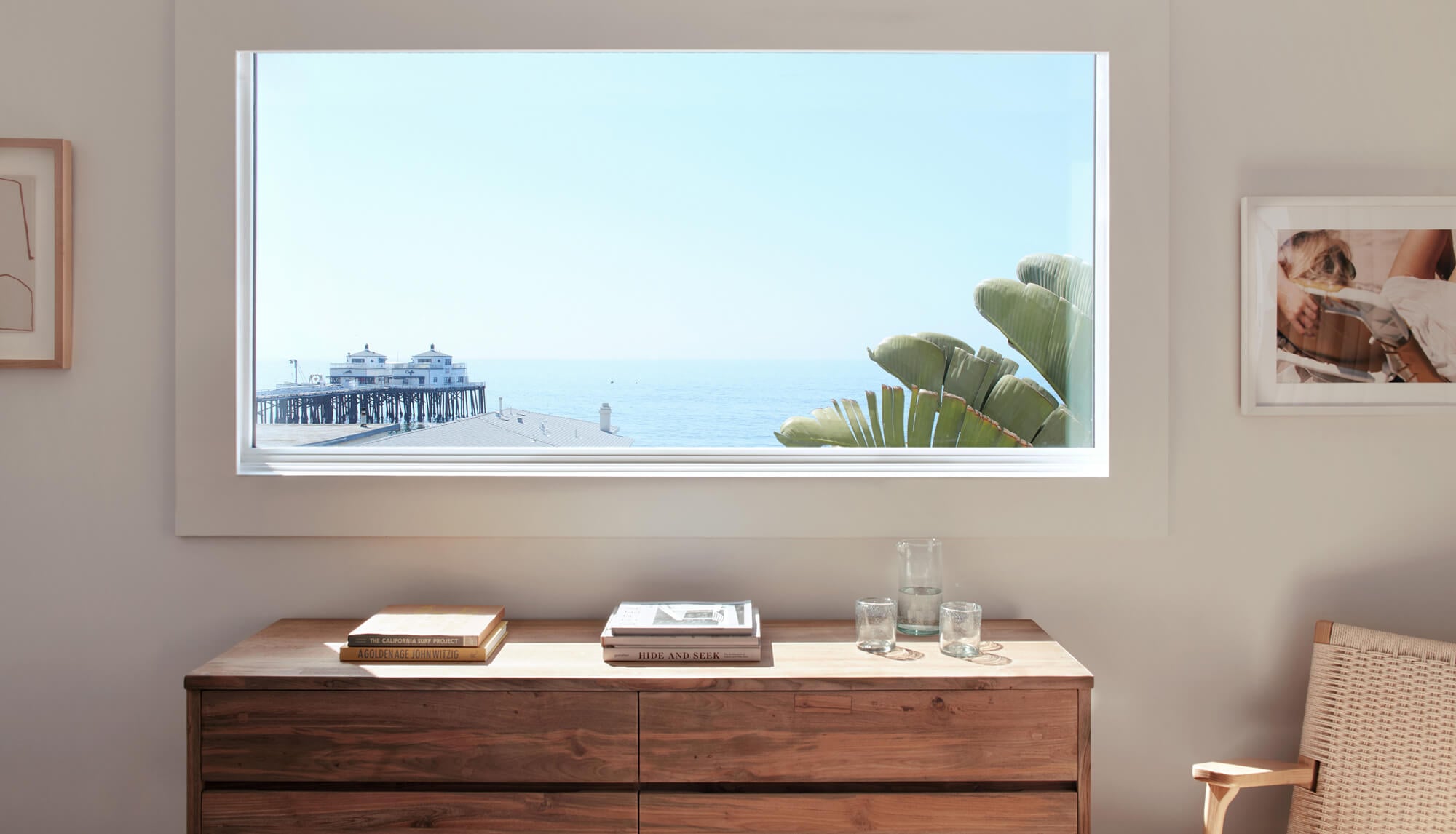 The Surfrider, North America — Coastal hotel room with panoramic window framing pier, ocean, and palm fronds above wooden dresser with books and glassware.