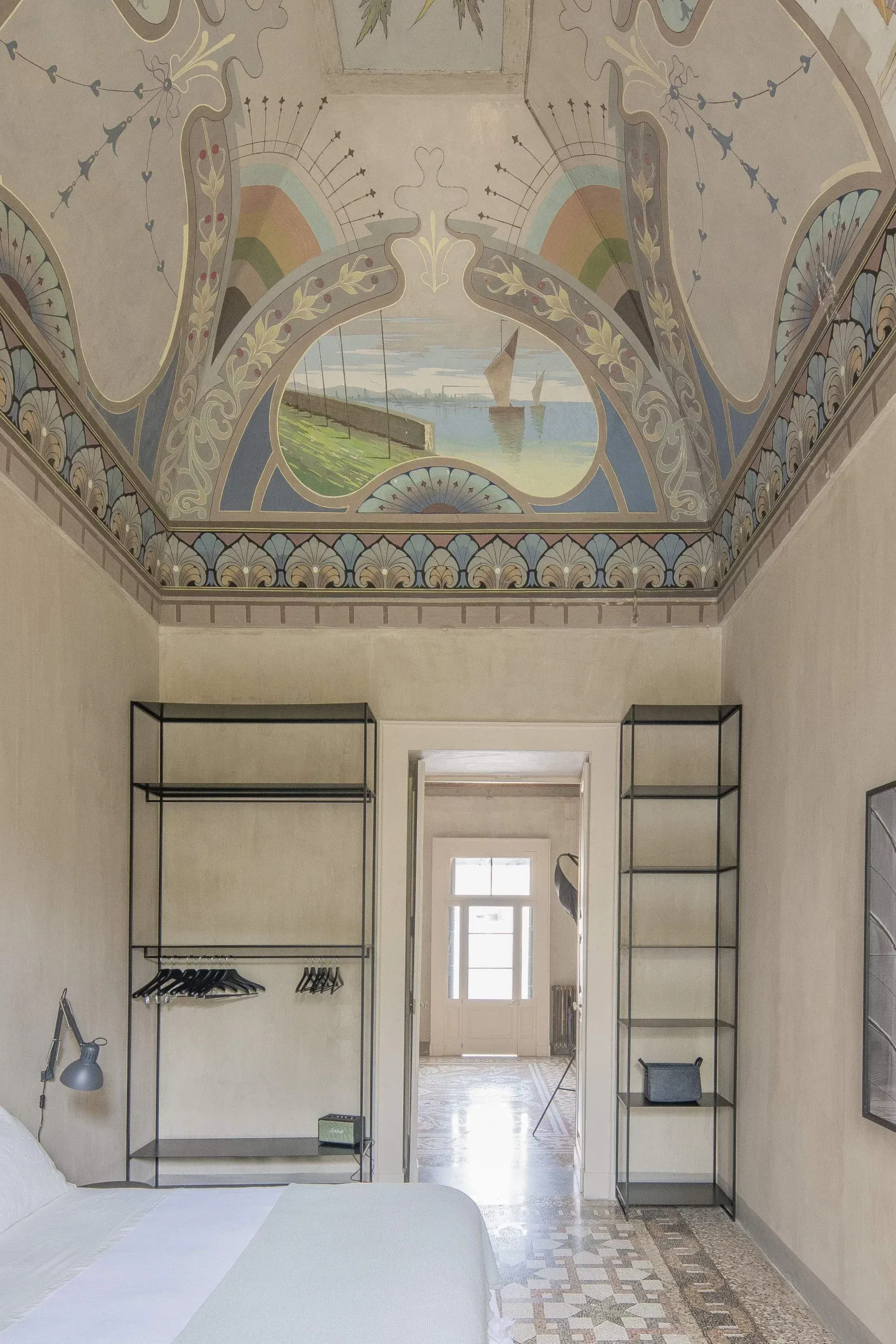 Palazzo Daniele, Europe — Bedroom with ornate frescoed ceiling, sailboat landscape mural, minimalist iron shelving, terracotta tile floor, serene neutral palette.